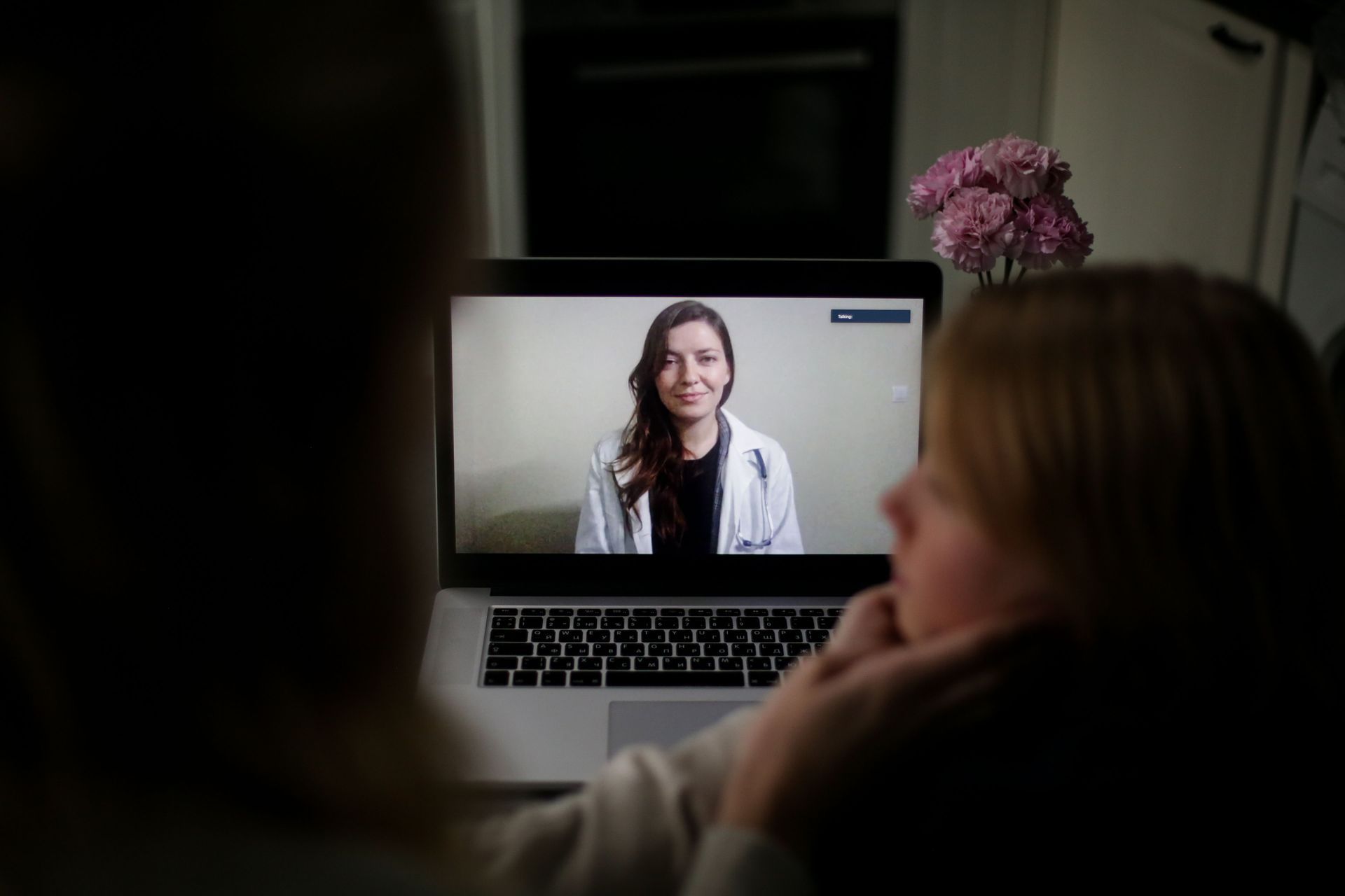 Person on laptop video call in a dim room, with two people watching from behind.