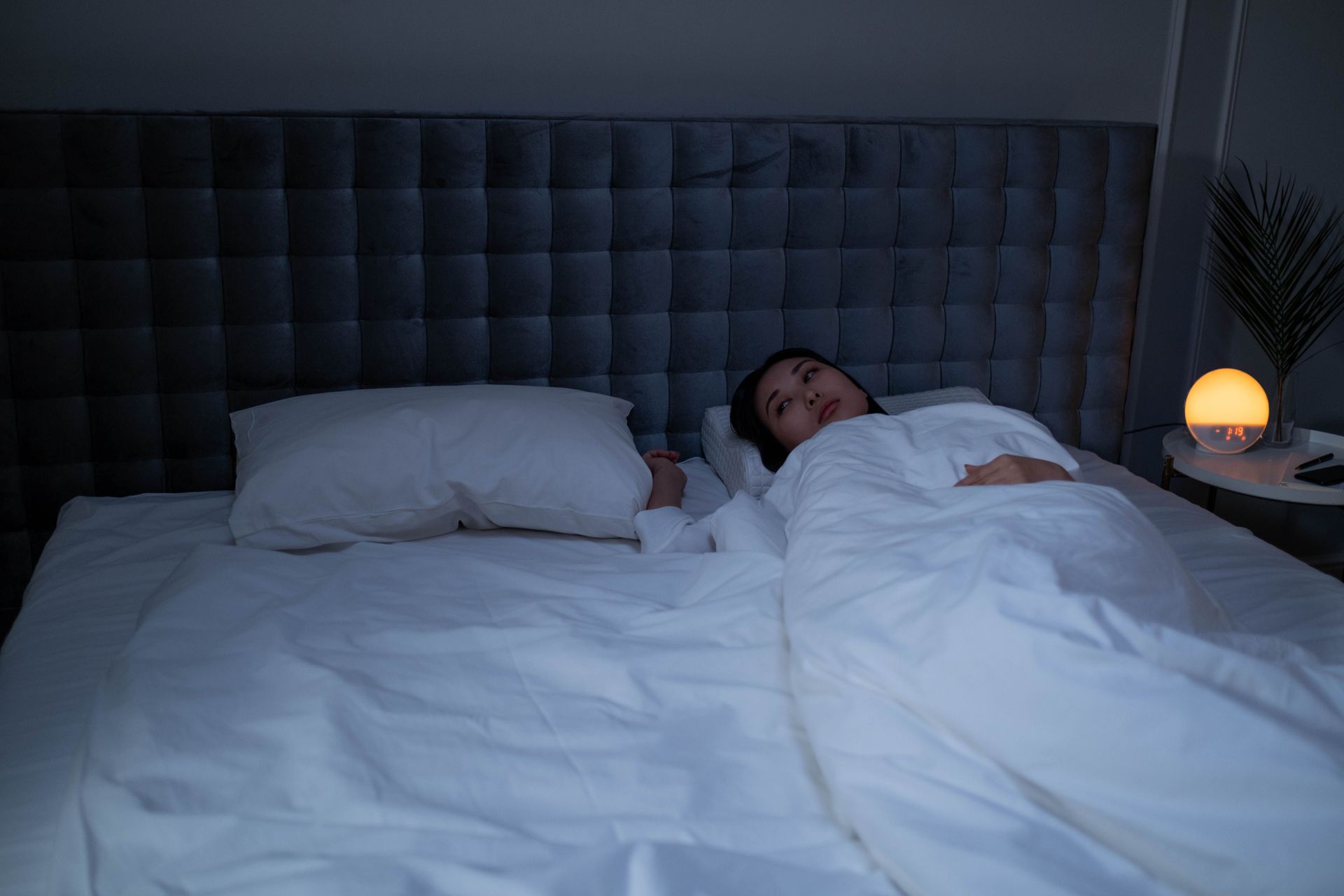 Person lying in bed under white covers beside an empty pillow, lit by a warm bedside lamp in a dark bedroom
