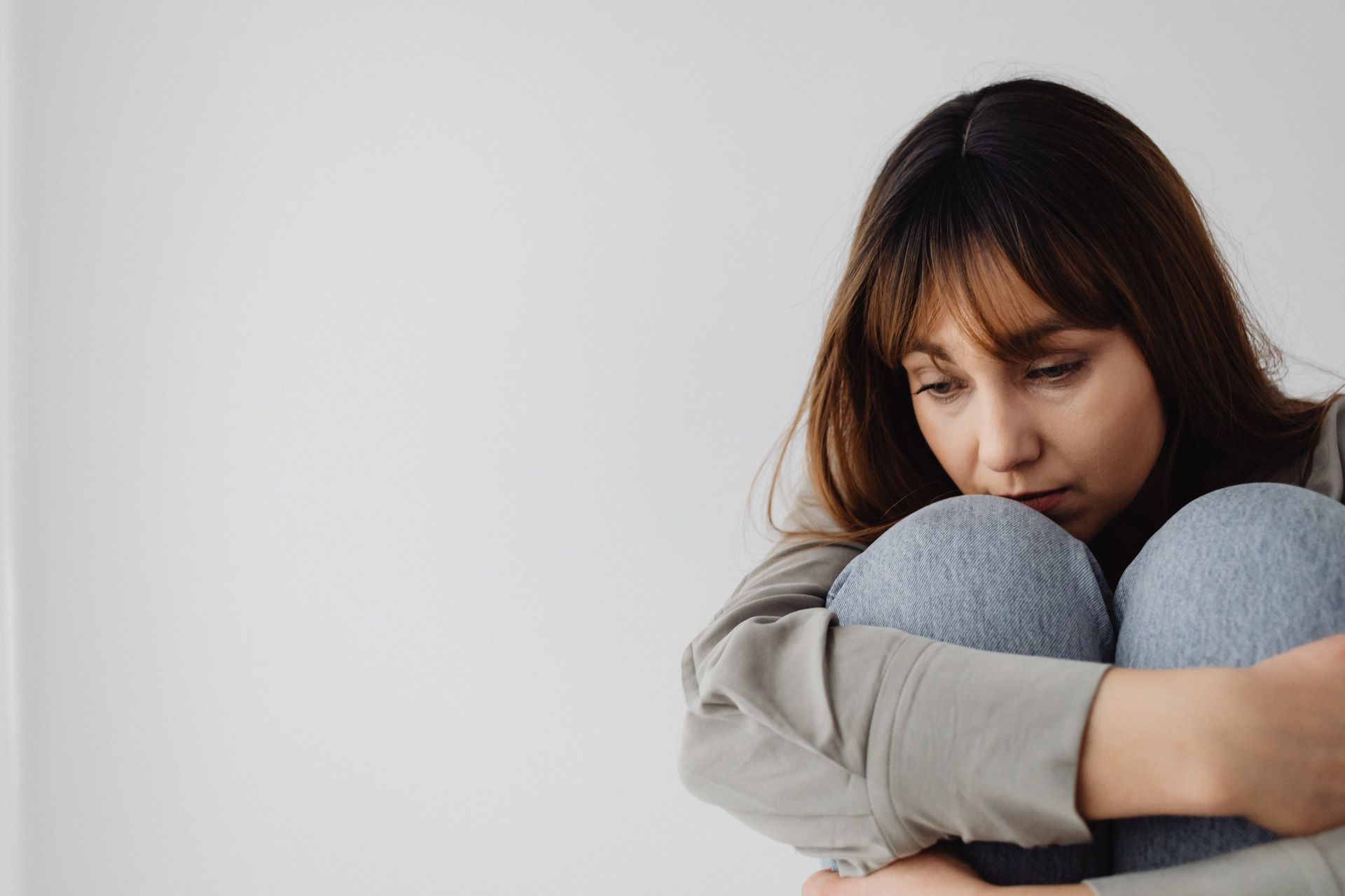 A distressed person sitting on the floor with knees hugged to chest, looking down in a gray room.