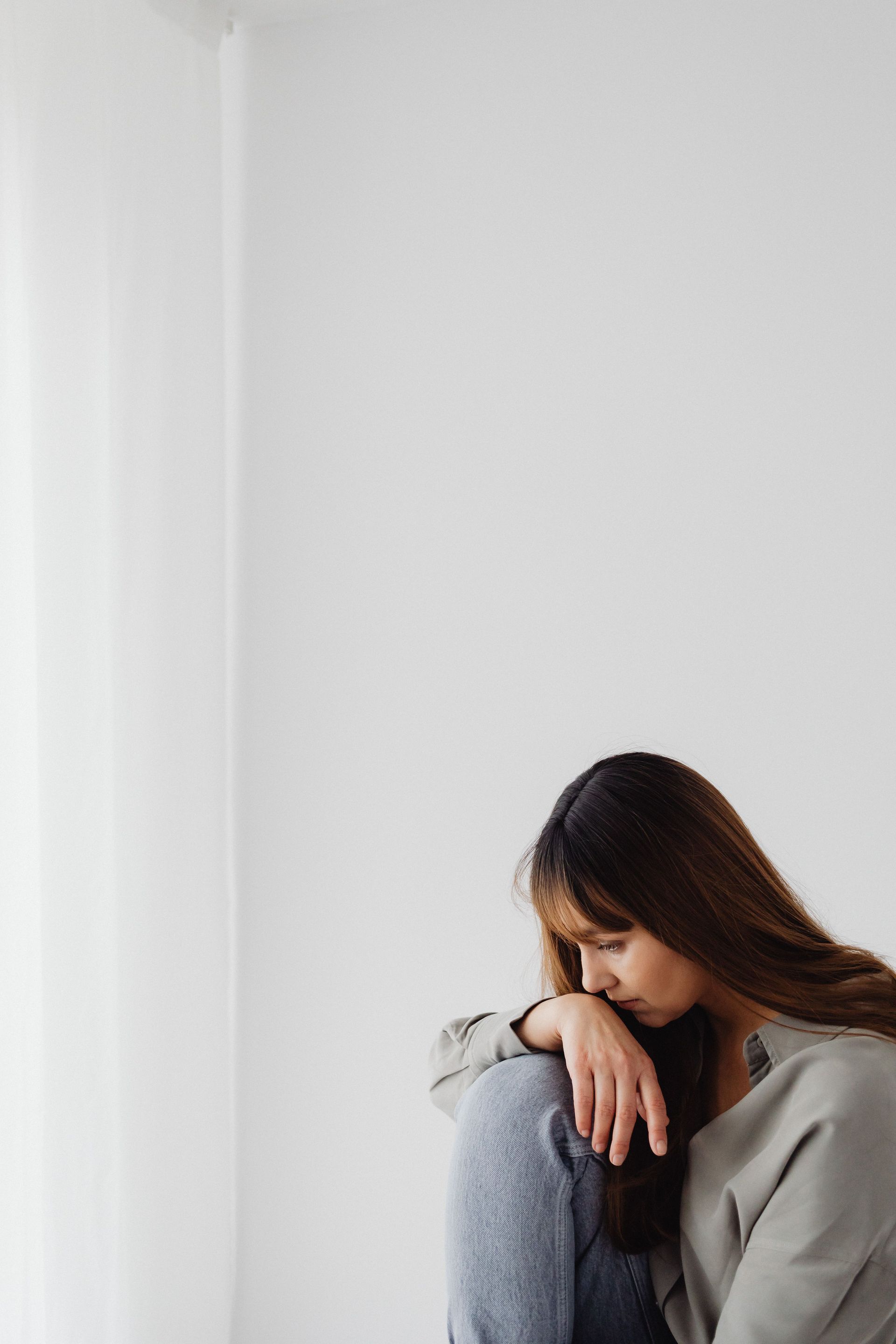 Person sitting by a white wall, resting head on folded arms with a pensive expression