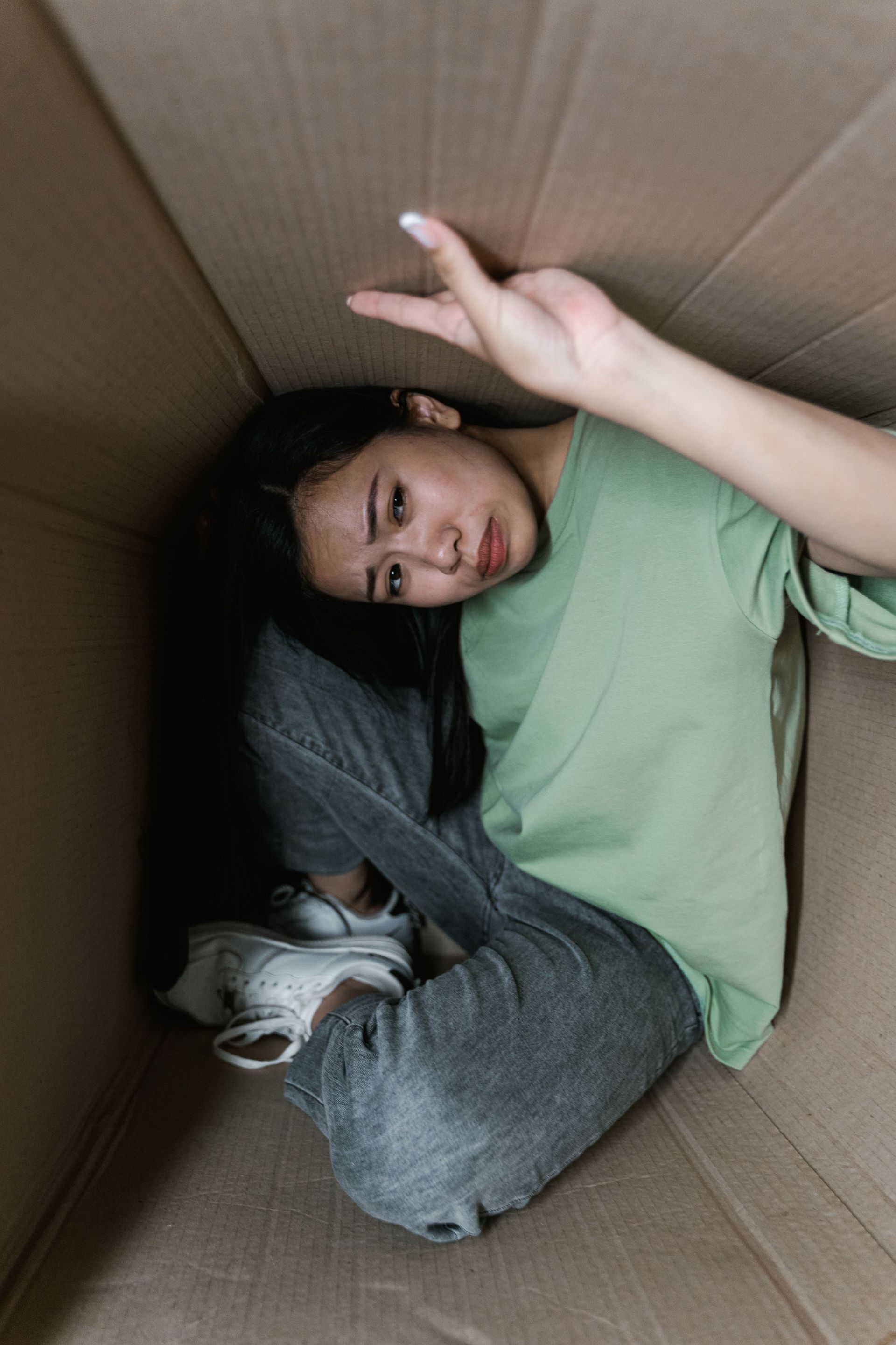 Person lying curled in a cardboard box, wearing a green shirt and gray pants, holding a peace sign
