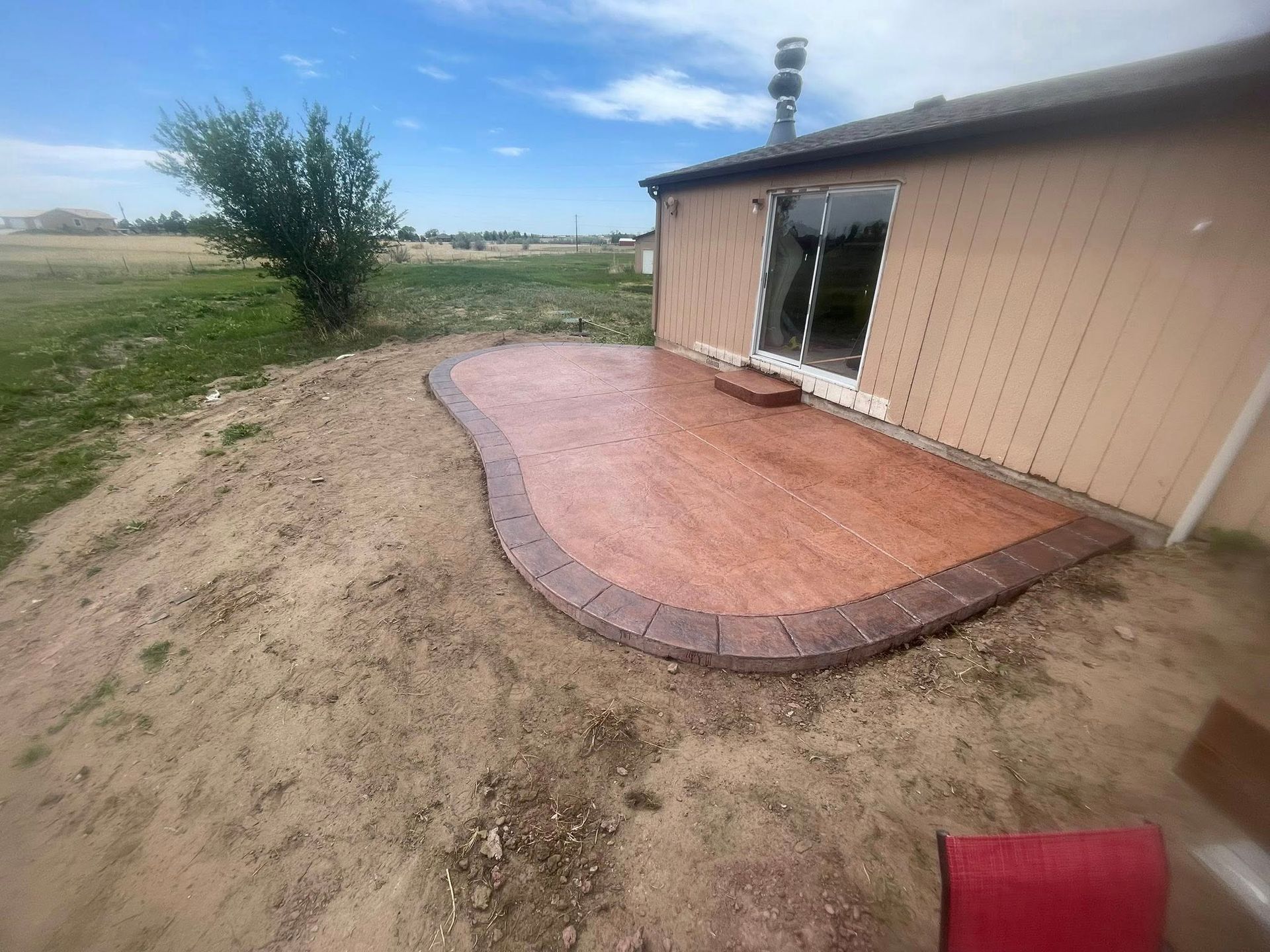 Curved brick patio beside tan building with open sliding door, on a dirt lot under a blue sky