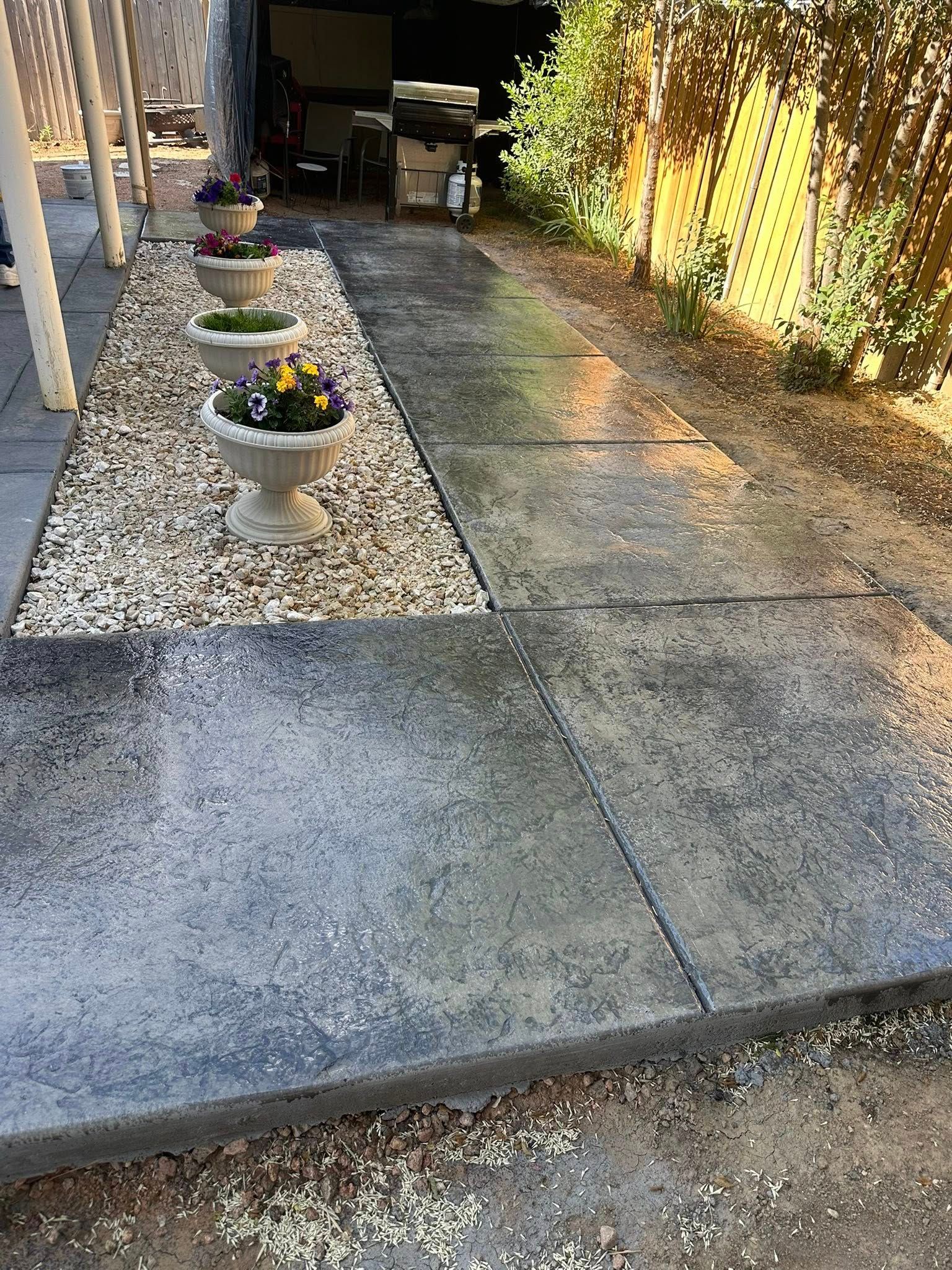 Concrete garden walkway beside potted flowers and a sunlit fence, leading to a shaded patio area