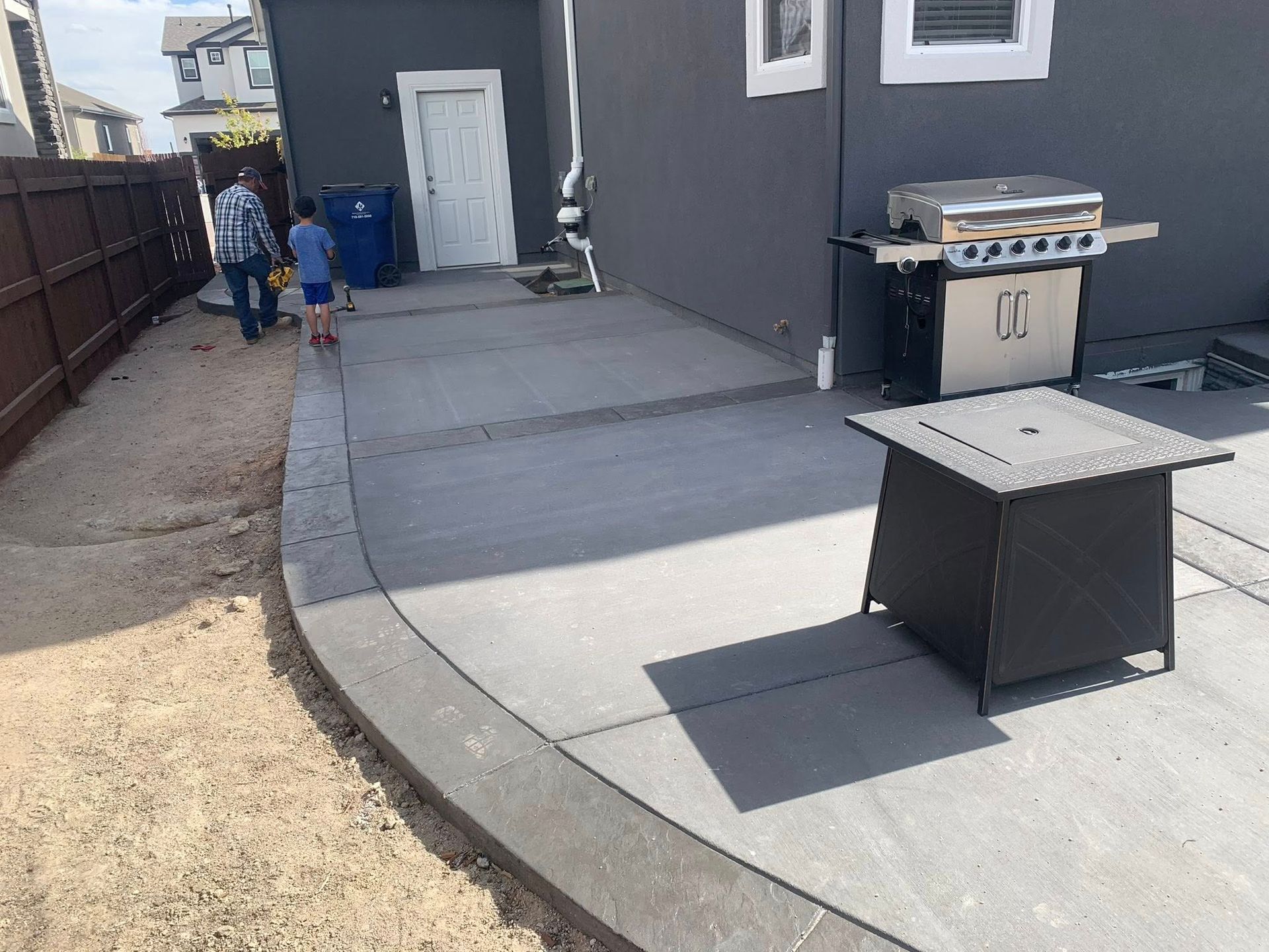 Backyard patio with grill, table, and a person near blue bins by a house wall