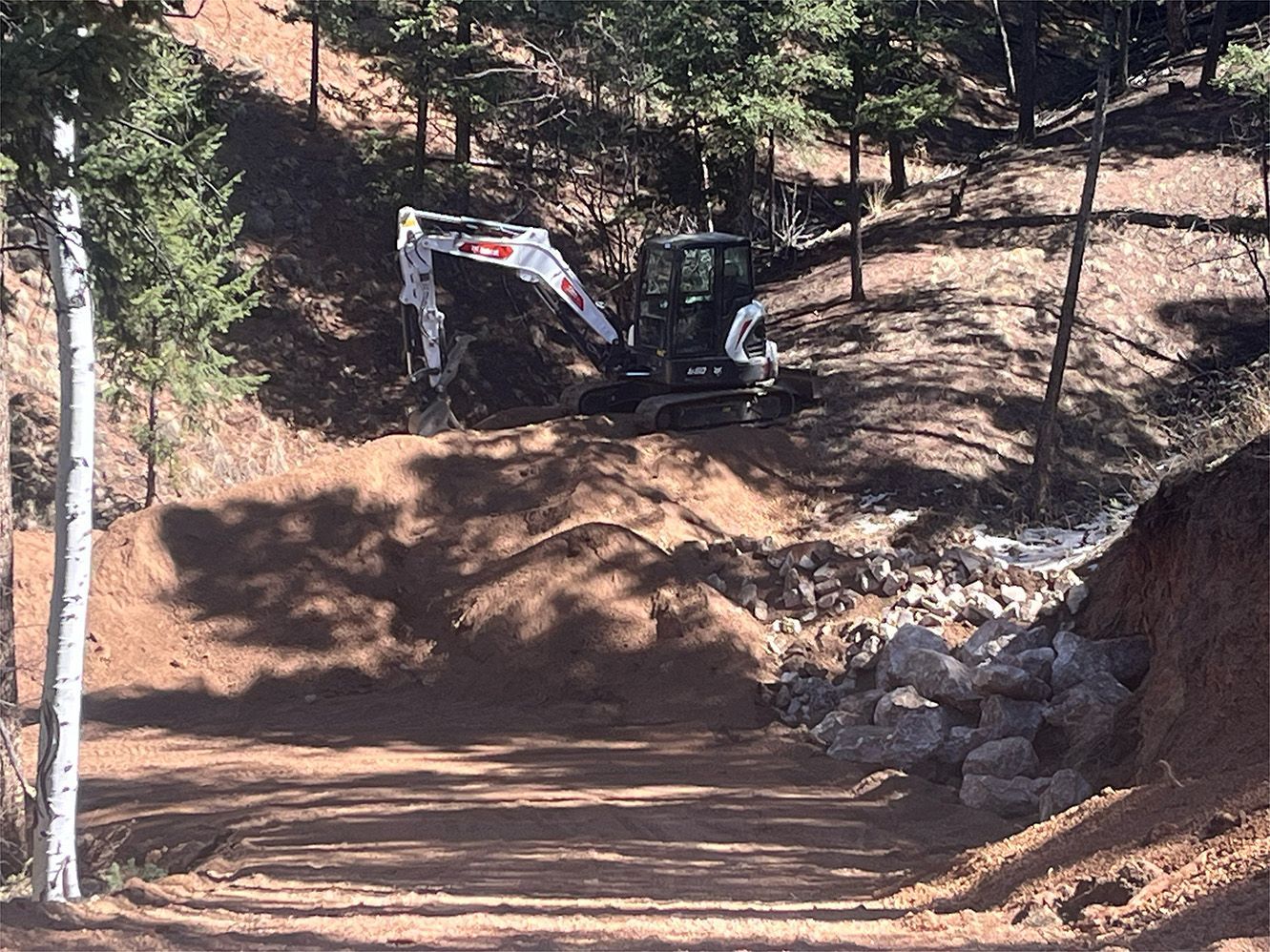 Excavator clearing dirt on a forested dirt road beside a rocky slope