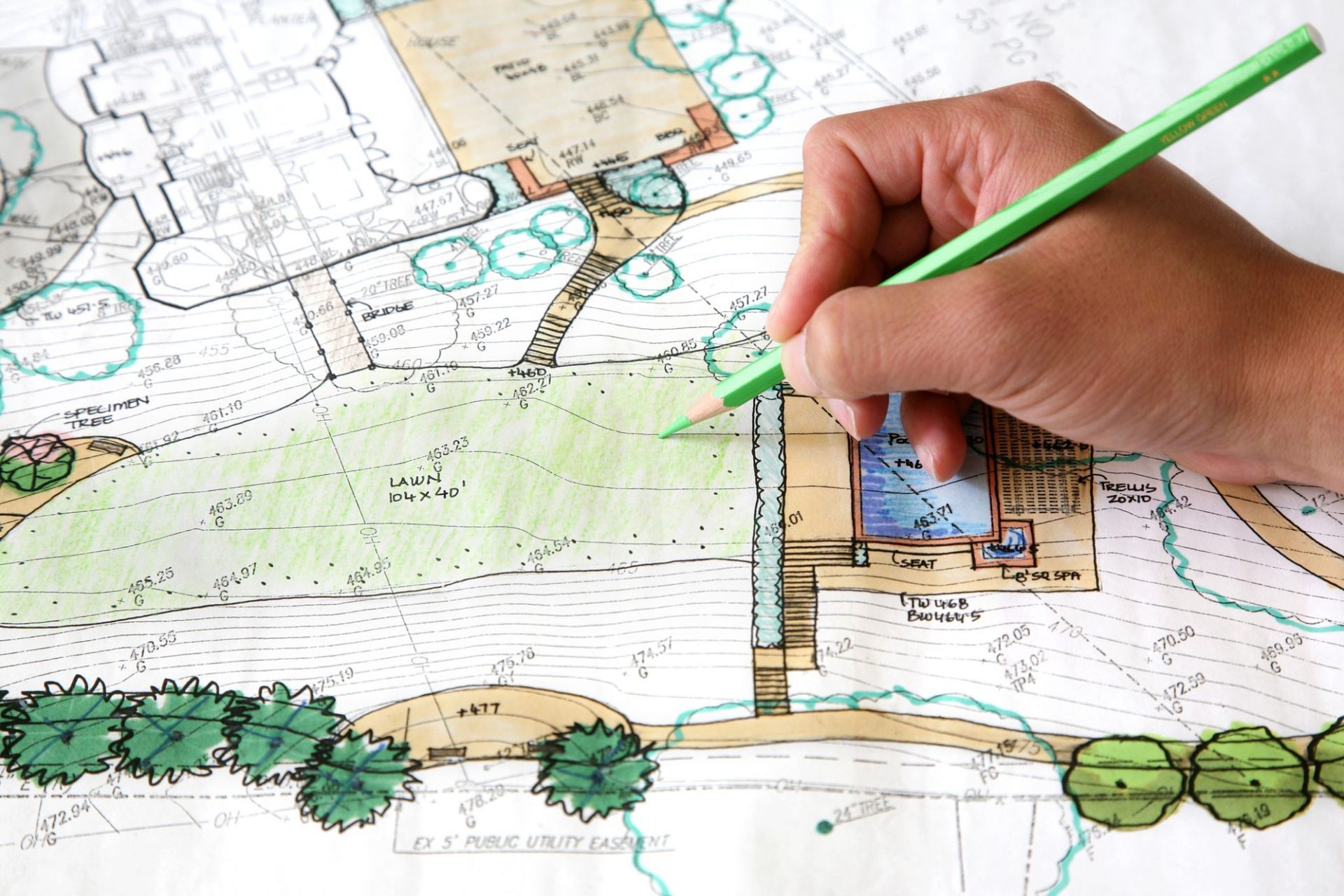 Hand with green pencil drawing on a landscape architecture plan, depicting a pool area, trees, and paths.