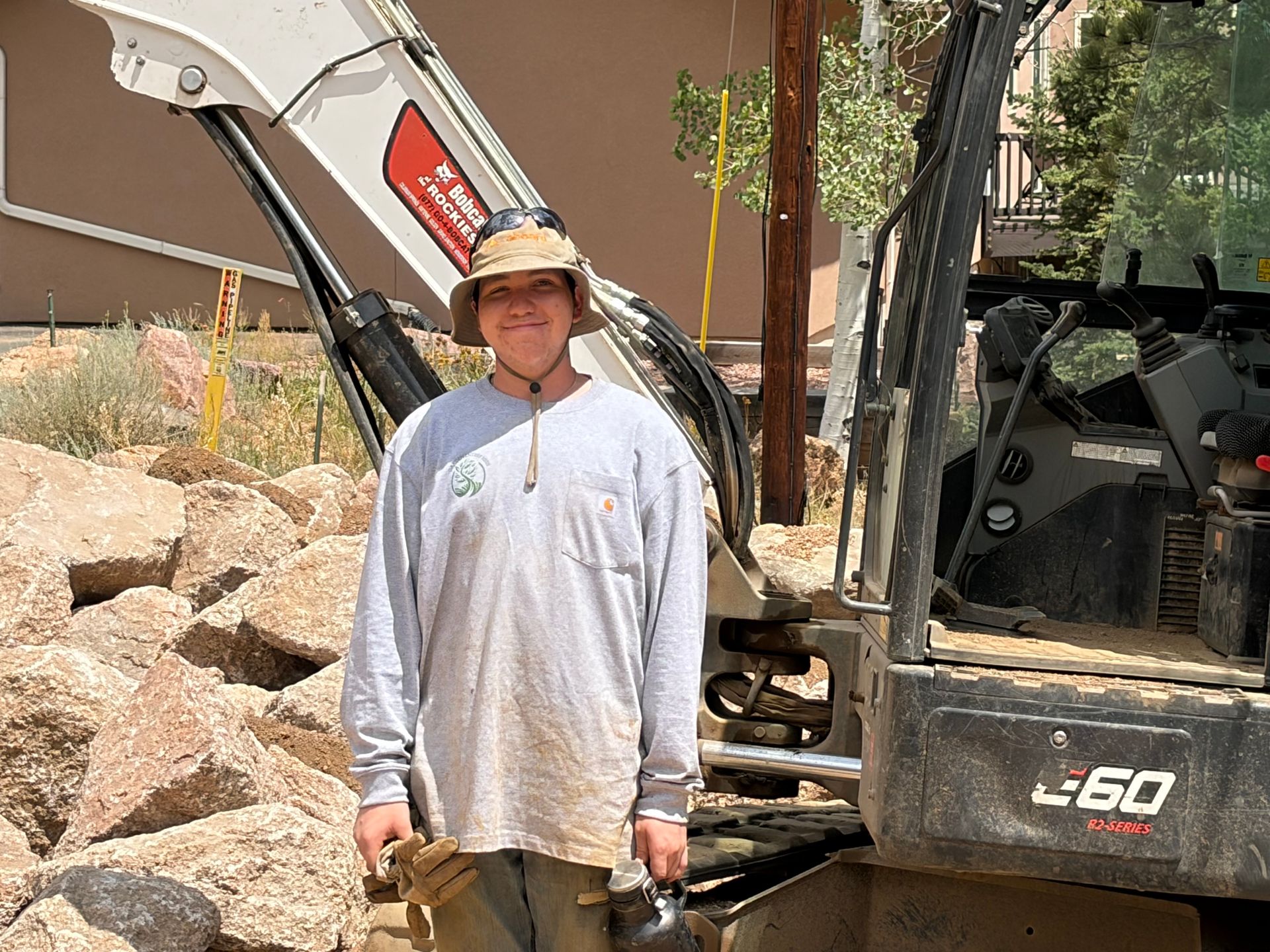Person standing beside a small excavator at a construction site, holding tools and wearing a hat.