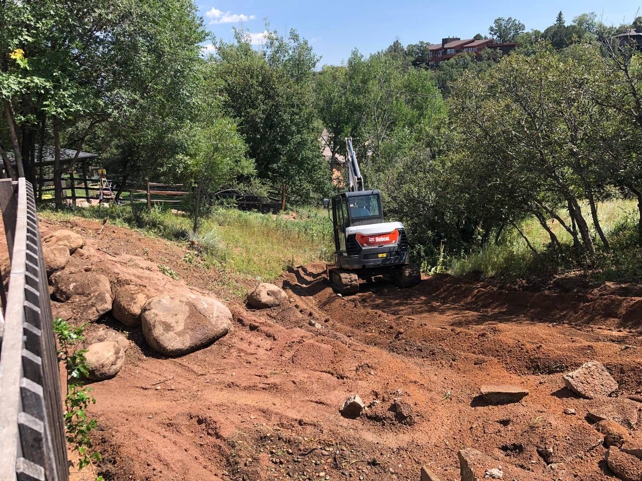 Small excavator clearing dirt in a wooded outdoor area beside a fence and large rocks.