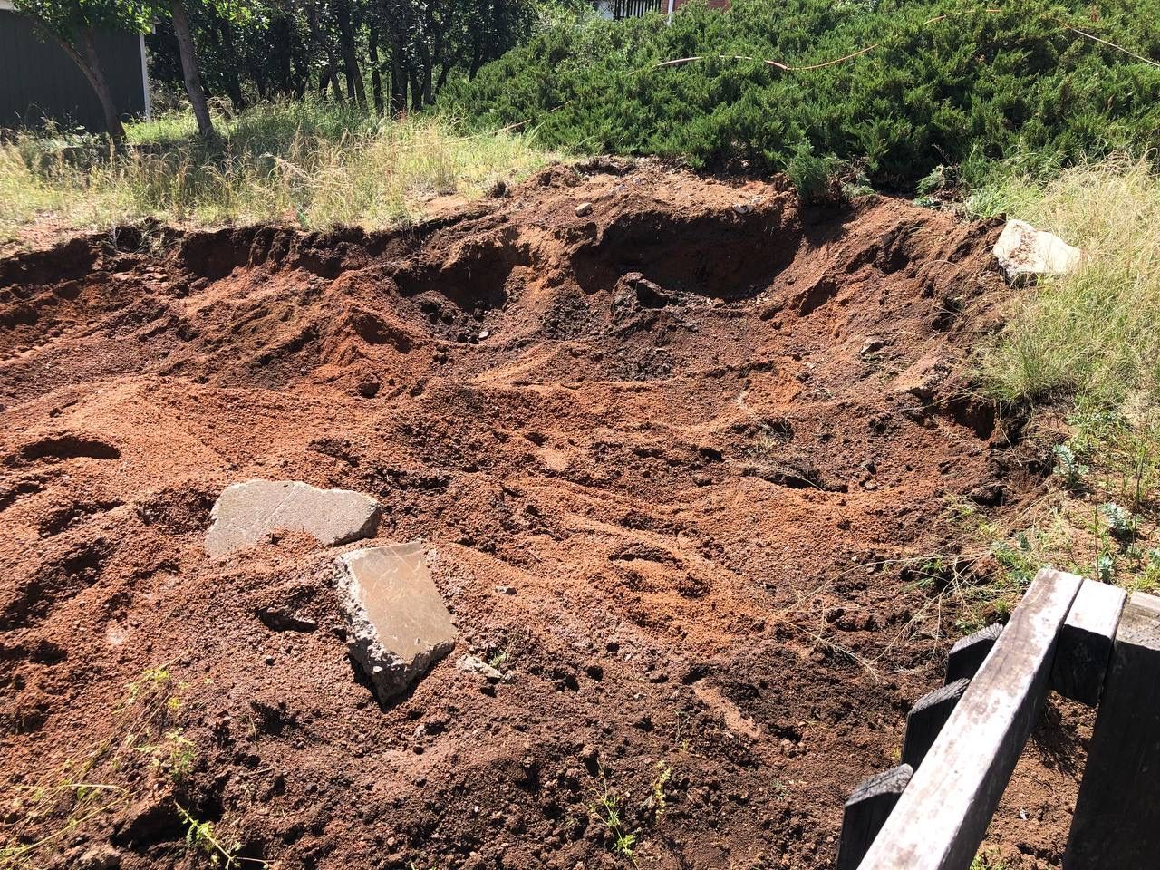 Large mound of reddish dirt in a grassy yard with scattered rocks and a shovel handle visible on the right