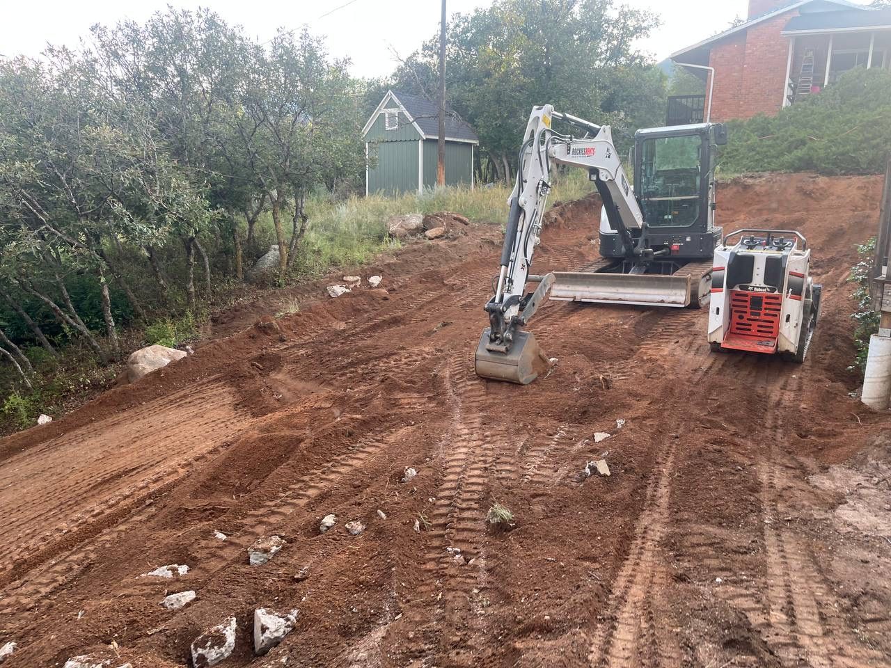 Excavator grading a muddy construction site beside bushes and a brick house.