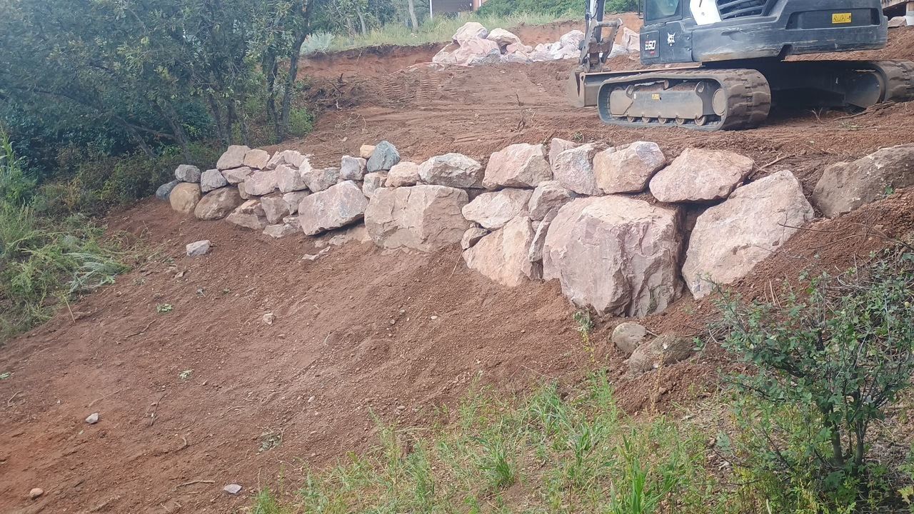 Excavator on a dirt slope beside a newly built stone retaining wall in a rural area