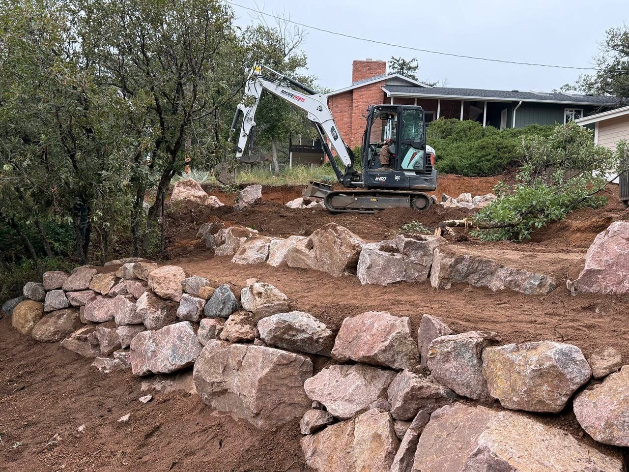 Excavator leveling a rocky backyard hillside beside a house and retaining wall under construction