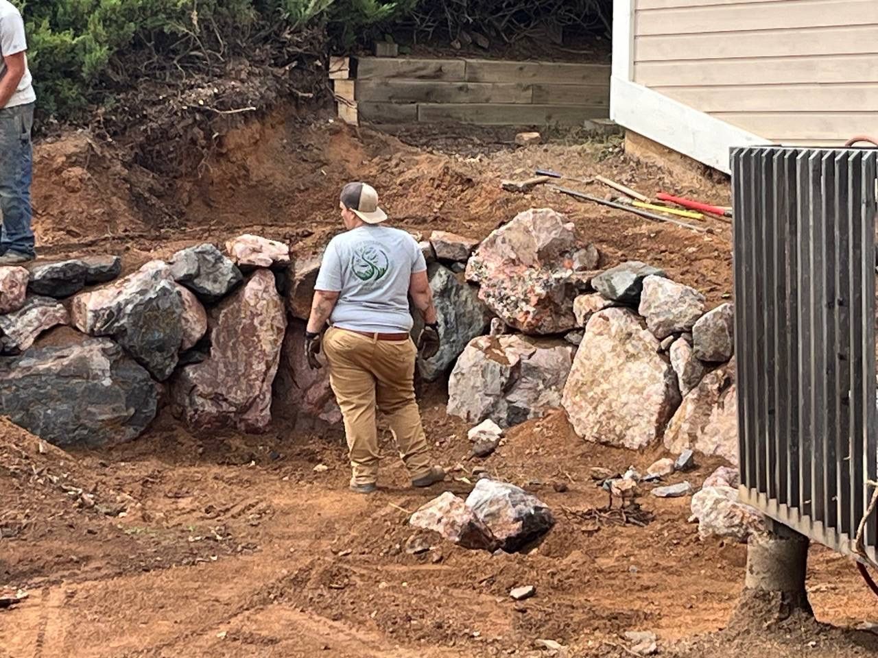 Worker arranging large rocks for a retaining wall in a dirt yard beside a house.
