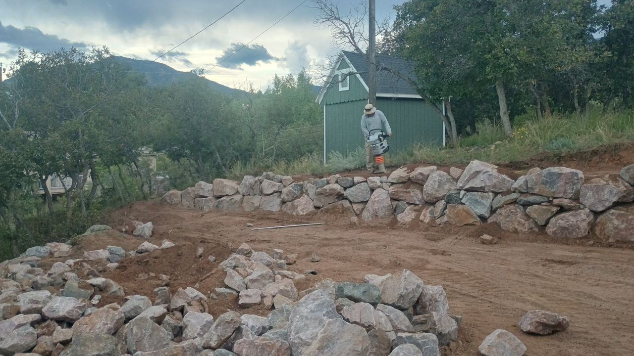 Rock-lined dirt path beside a green shed and trees, with mountains under a cloudy sky