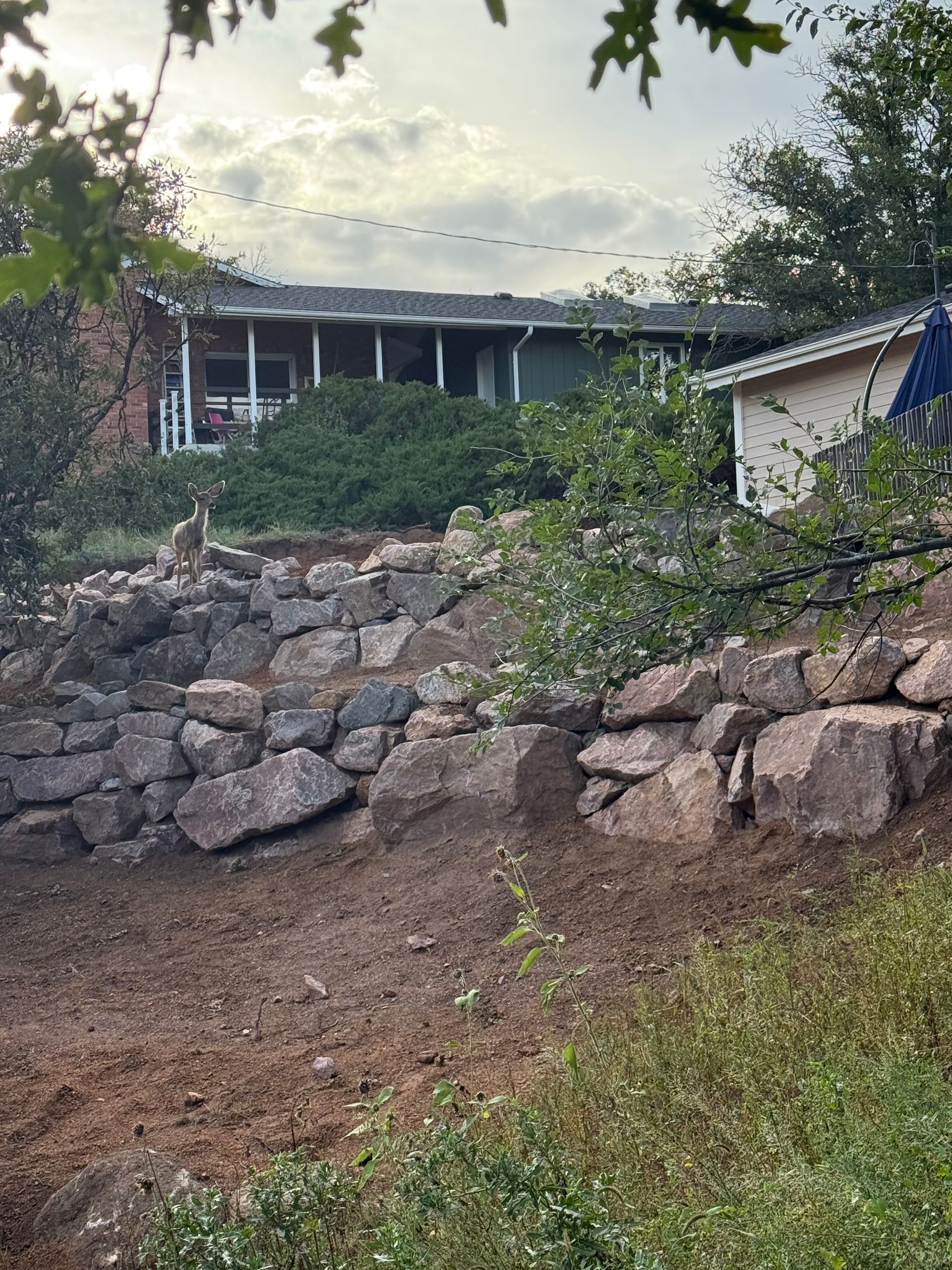 Rock retaining wall and dirt path leading up to a house with trees and cloudy sky.