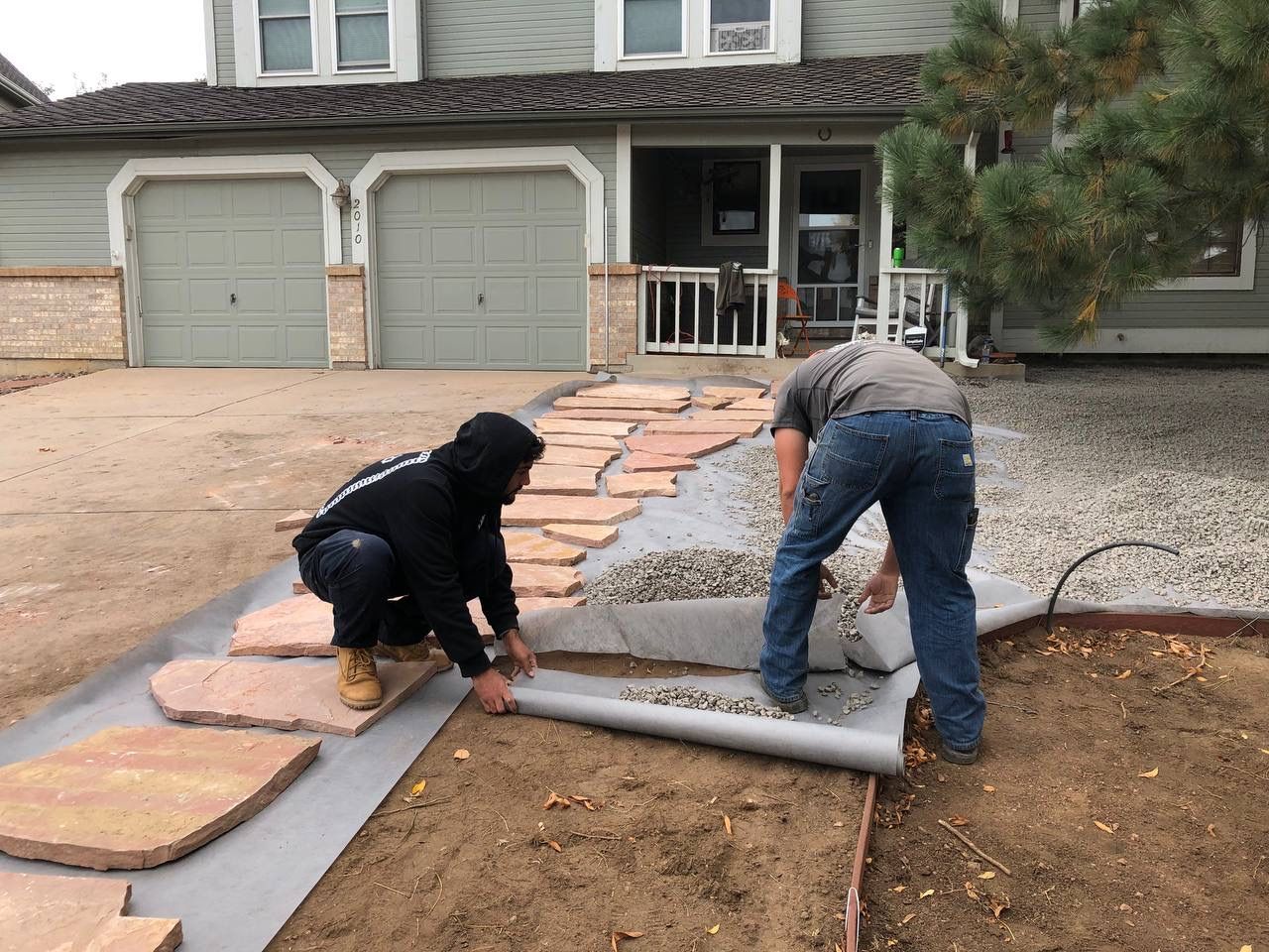 Two workers laying a stone walkway beside a house driveway and garage.
