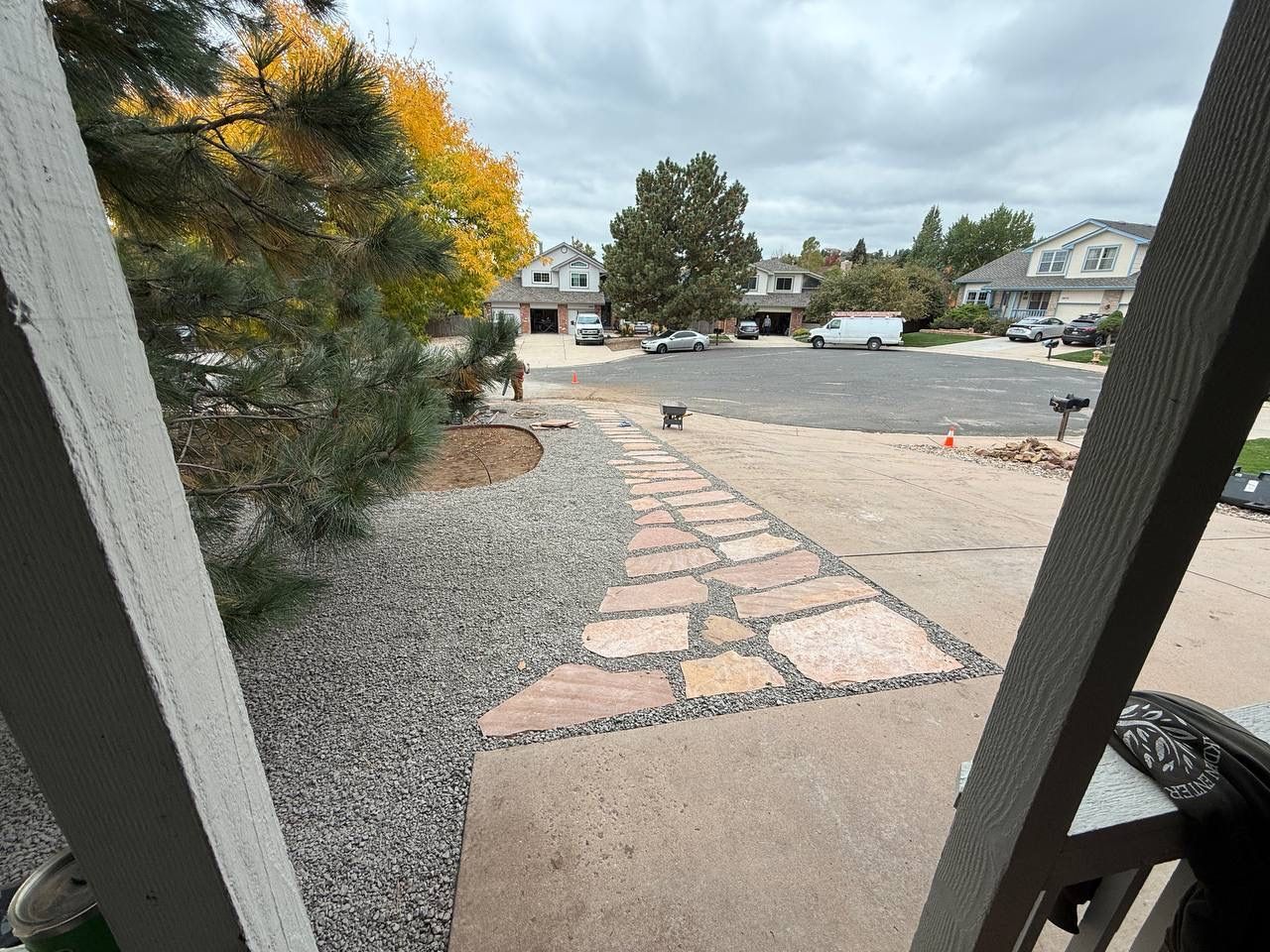 Front porch view of a suburban street with a stone walkway, gravel, trees, and parked cars