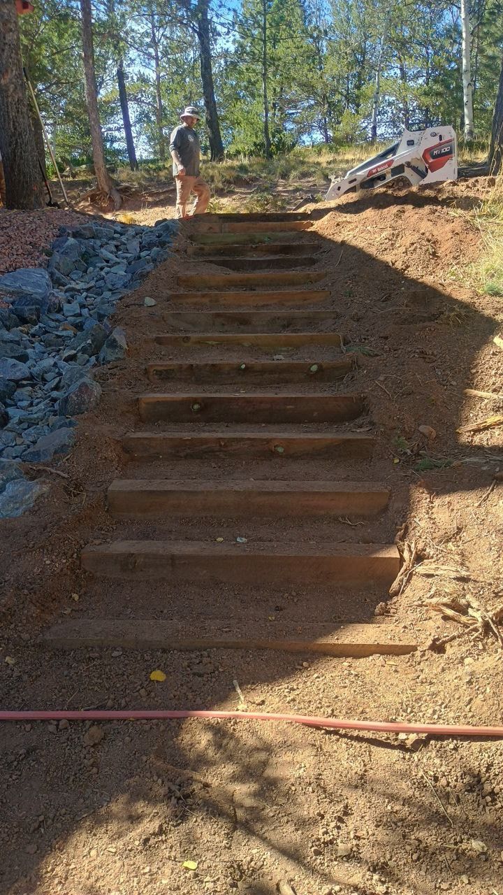 Steep dirt staircase under construction on a wooded hillside, with a worker at the top and gravel on the left