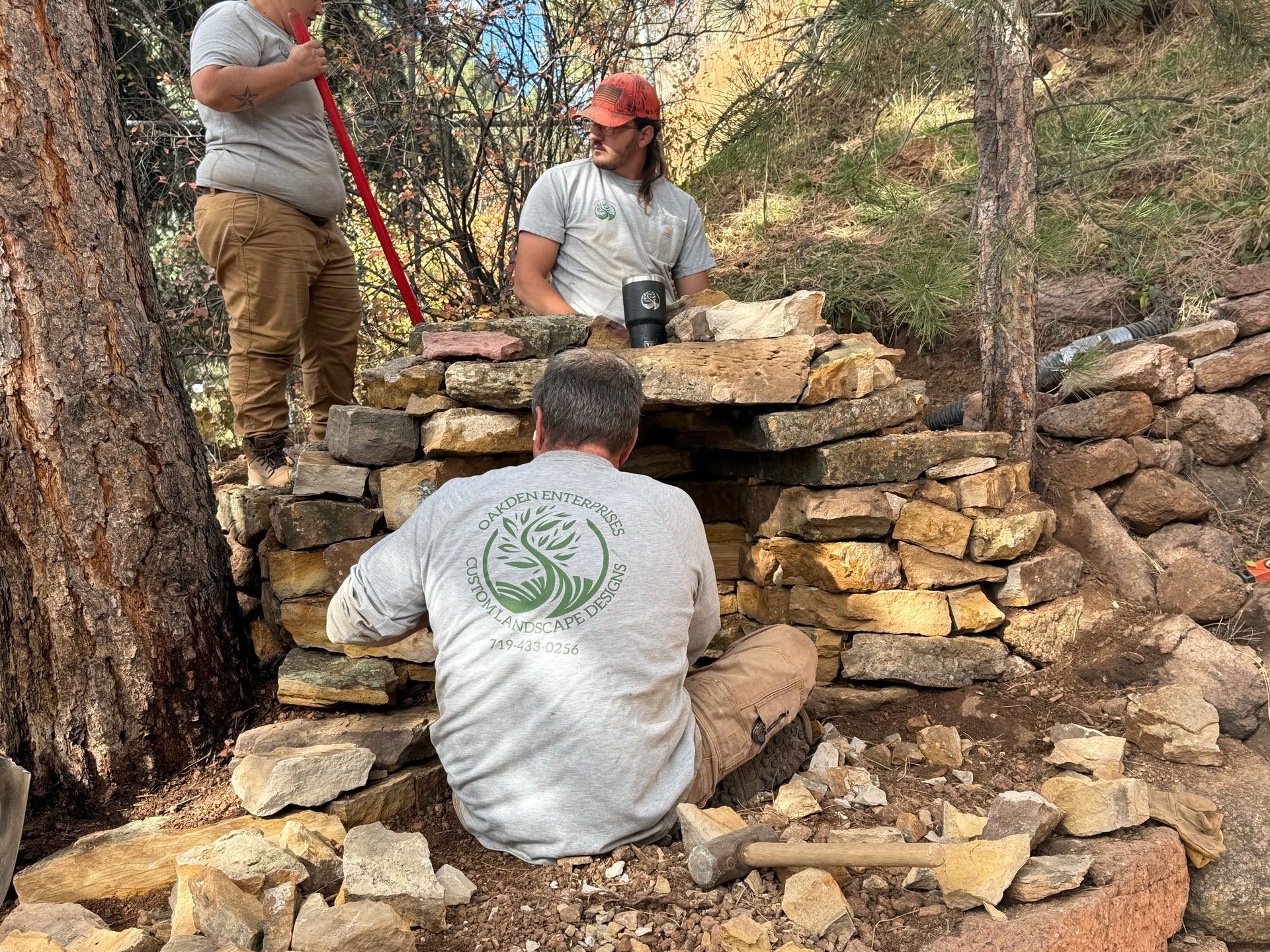 Three people stacking a dry stone wall in a wooded outdoor area
