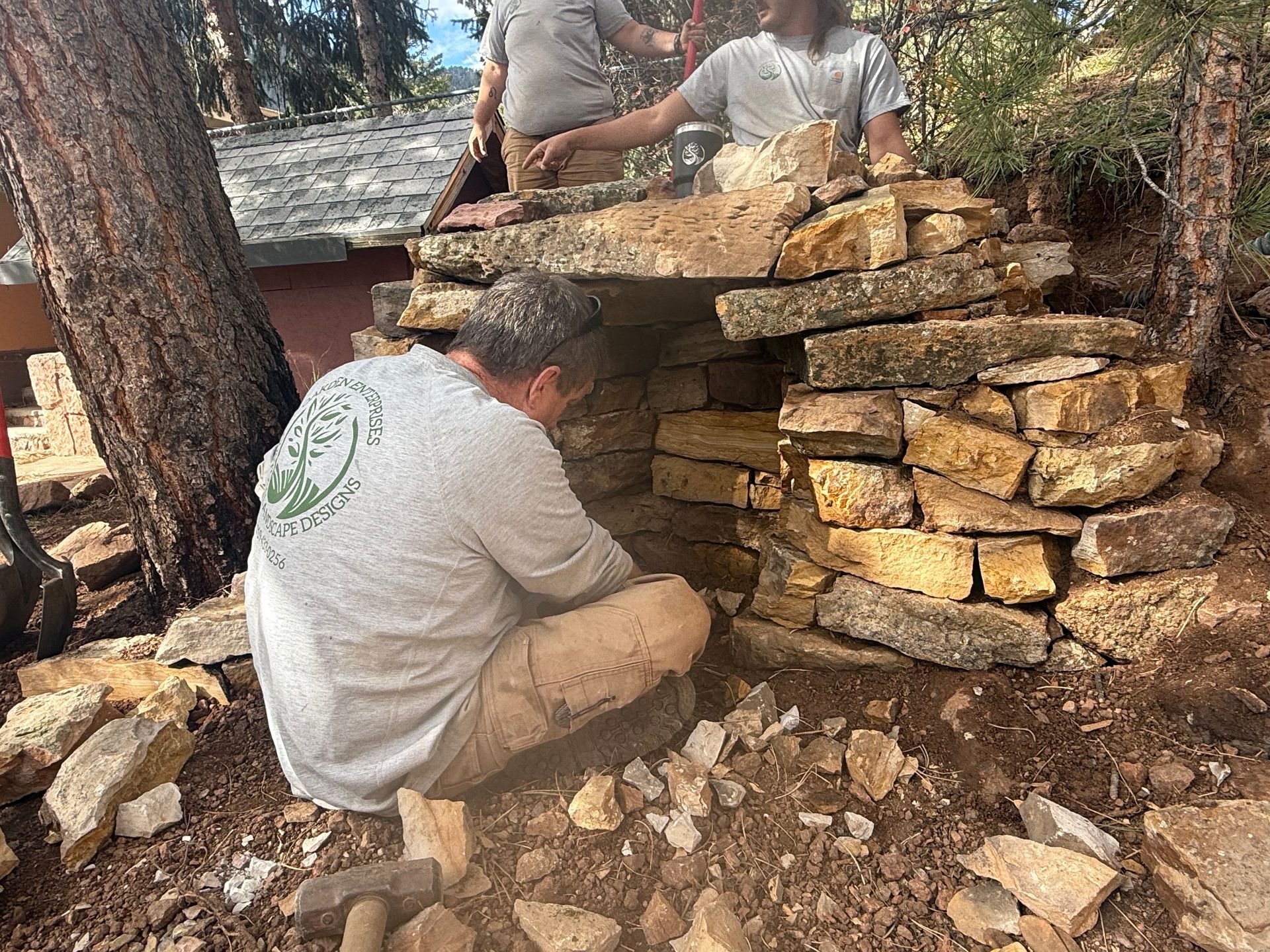 Two people building a stacked stone wall outdoors in a wooded area