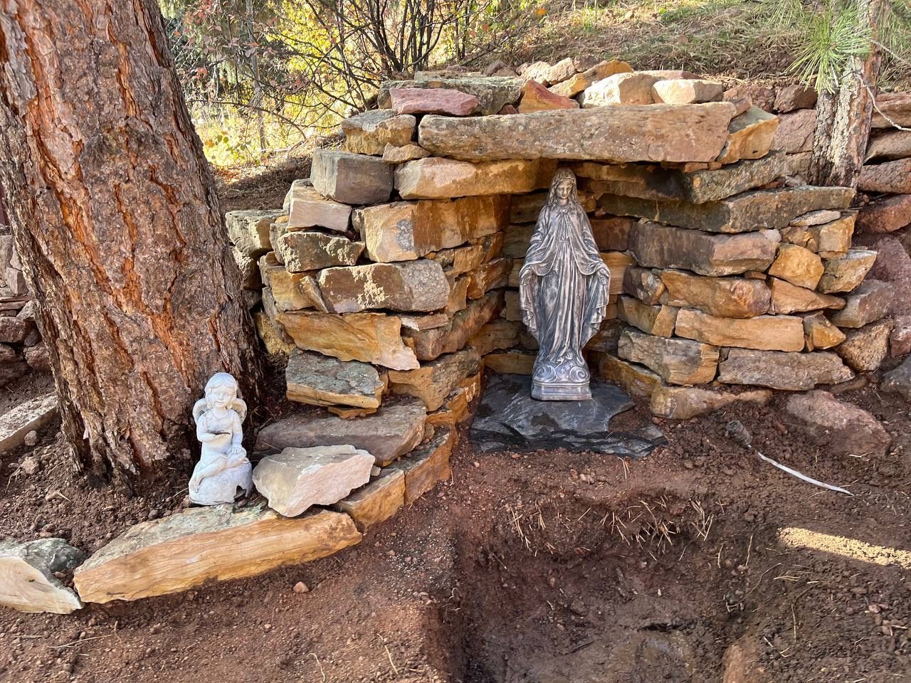 Stone shrine alcove with two small statues beneath a tree in a rocky outdoor setting