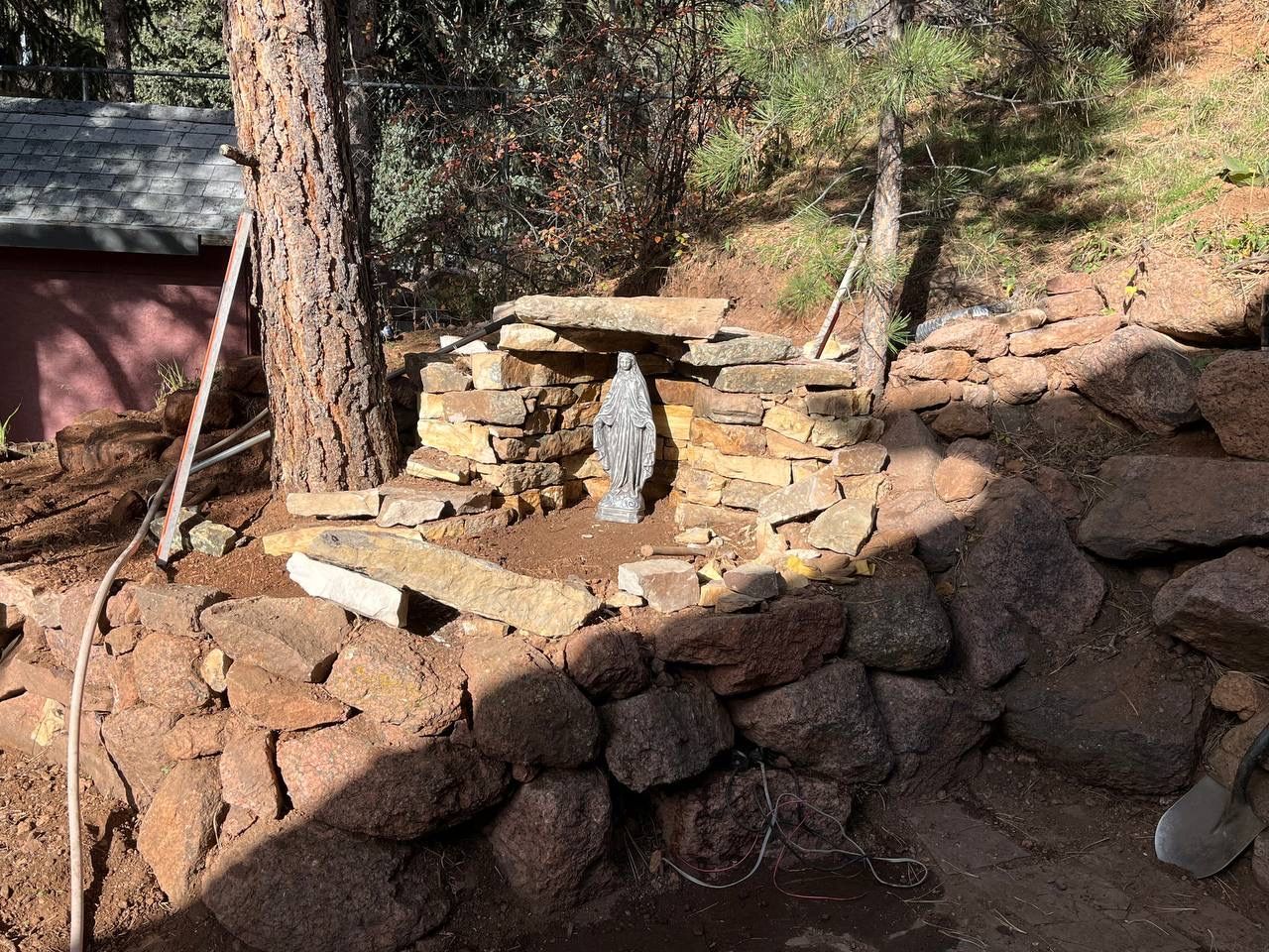 Small outdoor stone shrine with a statue, built into a rocky hillside among trees.