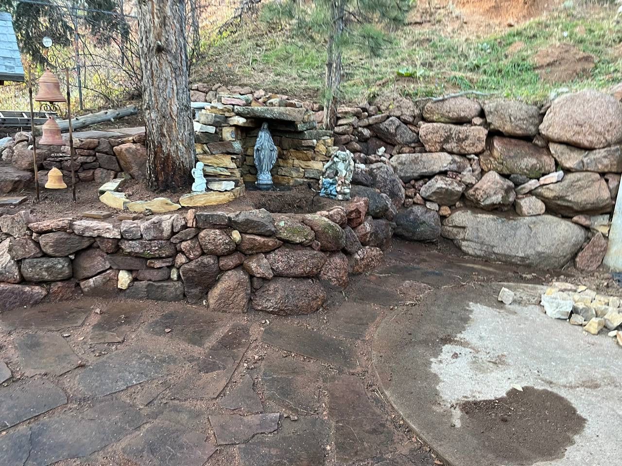 Stone garden shrine with small blue statues beside a tree and a rocky retaining wall