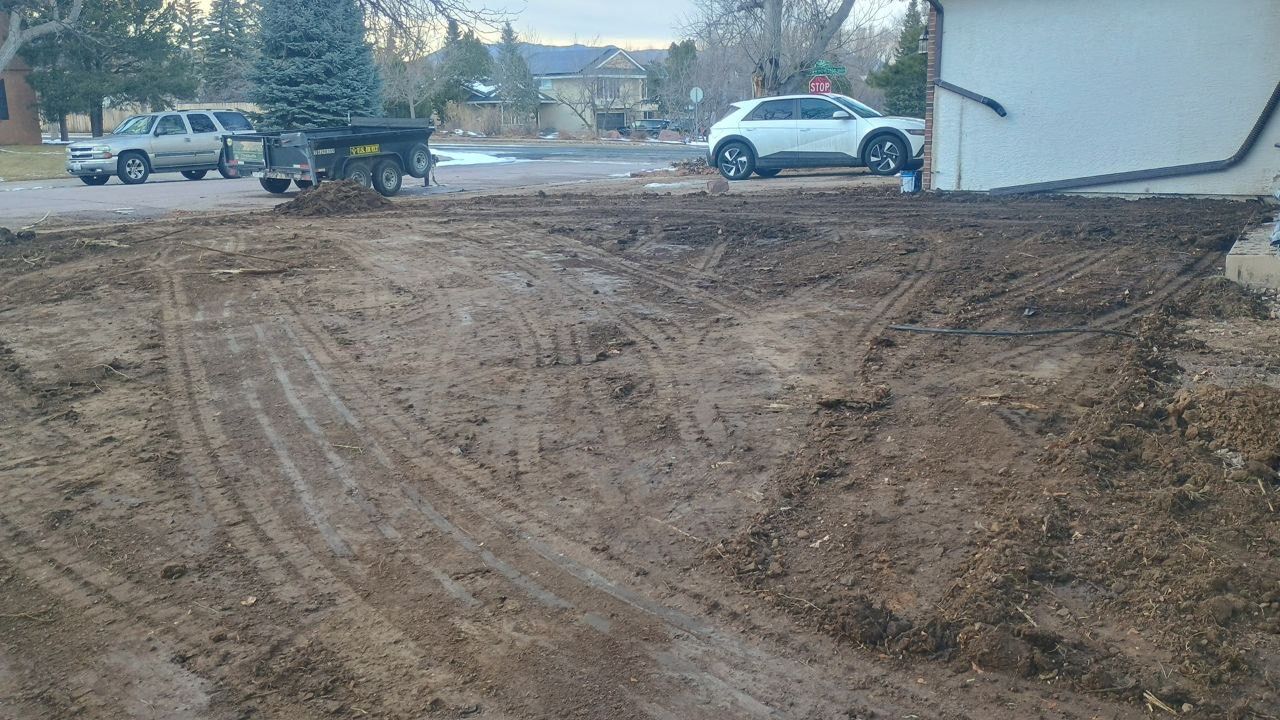 Mud-covered driveway beside a house, with parked cars and a trailer in the background.