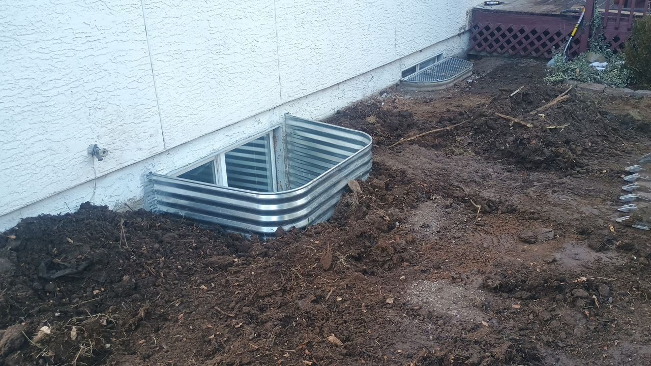 Stacked metal window wells beside a house foundation on bare dirt and gravel.