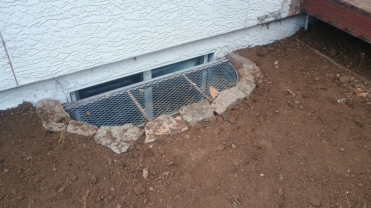 Small basement window well with metal grate beside a white house wall and dirt ground