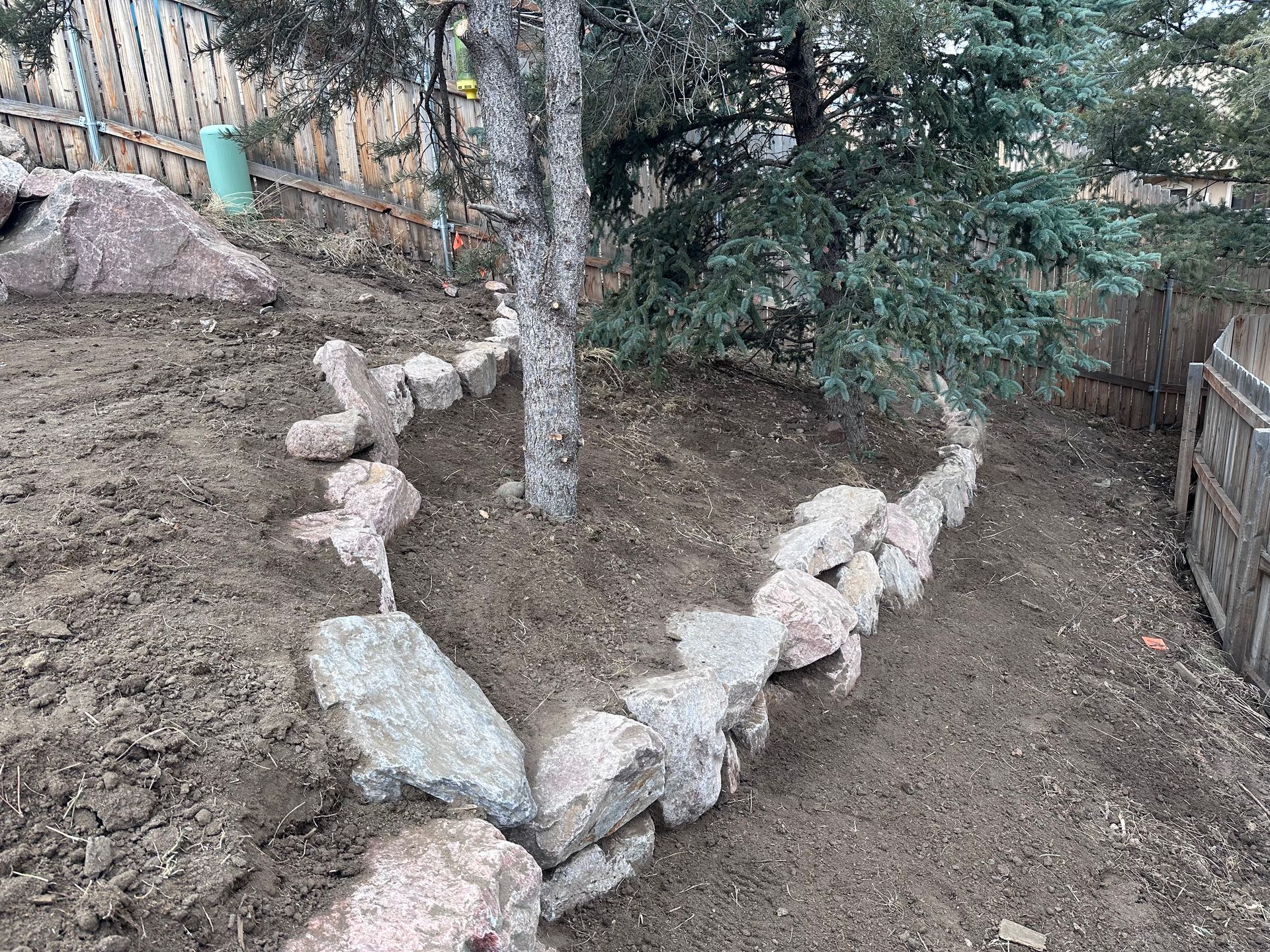 Rock-bordered dirt yard with a tree in the center and a wooden fence on the right