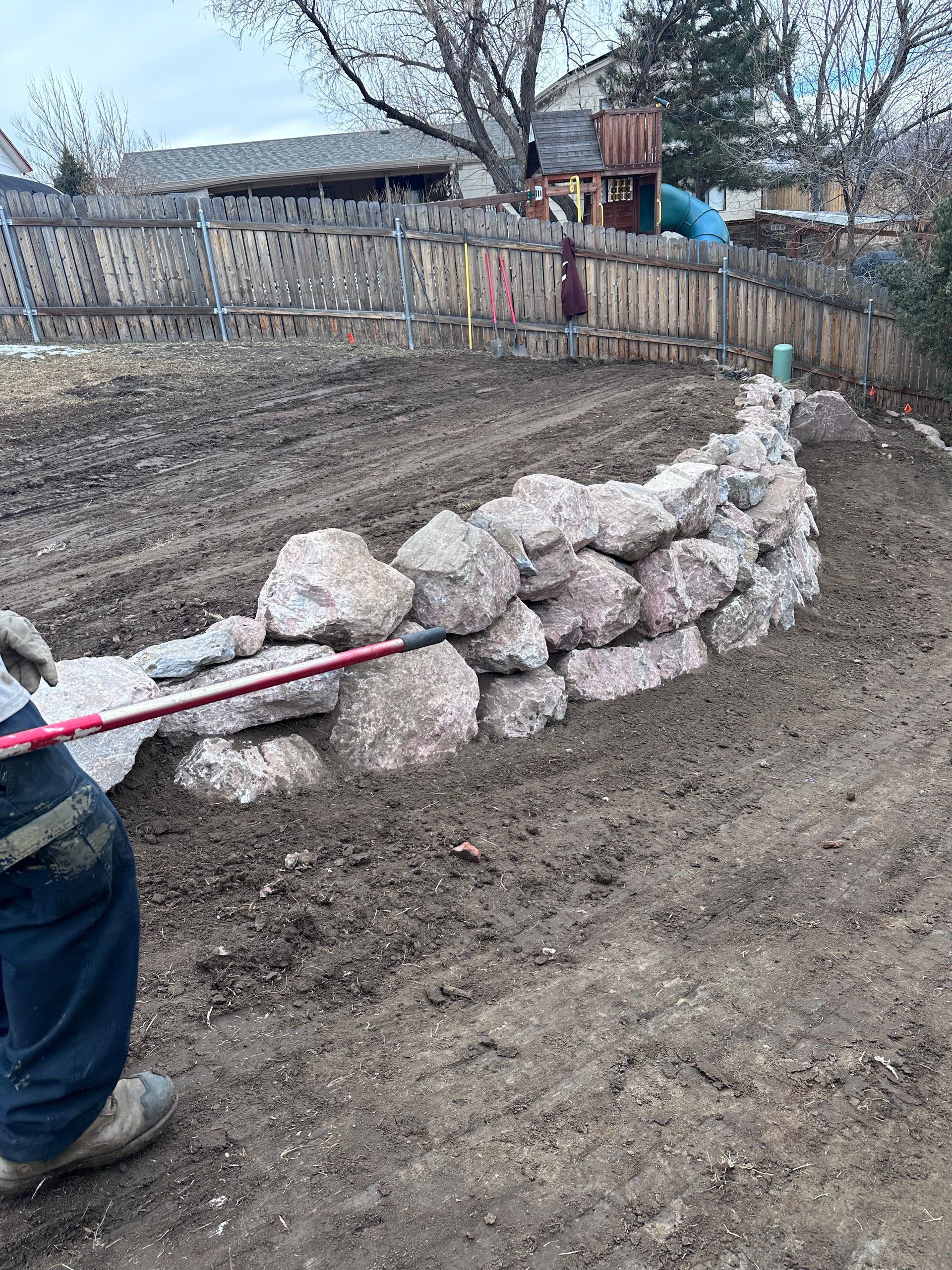 A person in a yard pulls a red rope attached to a low stone-lined trench near a fence.