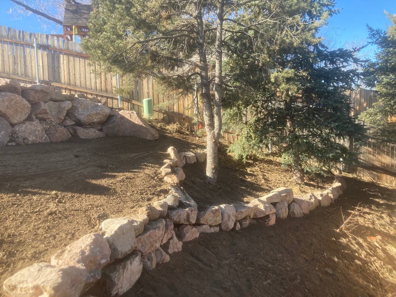 Rocky landscaped yard with a tree, stone retaining walls, and a wooden fence in the background