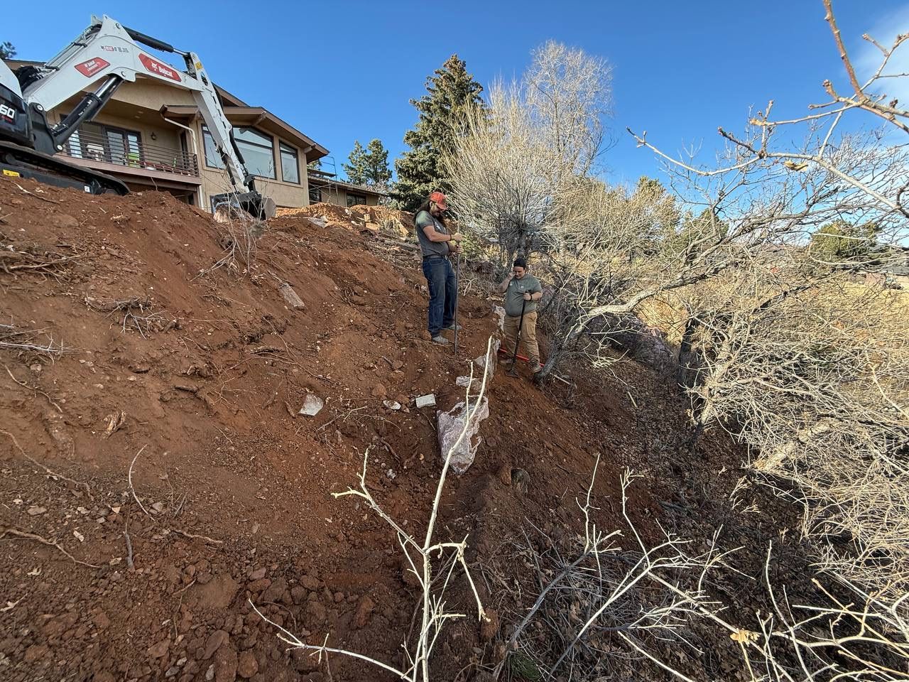 Worker stands on a steep dirt slope beside a house with exposed foundation and fallen trees.