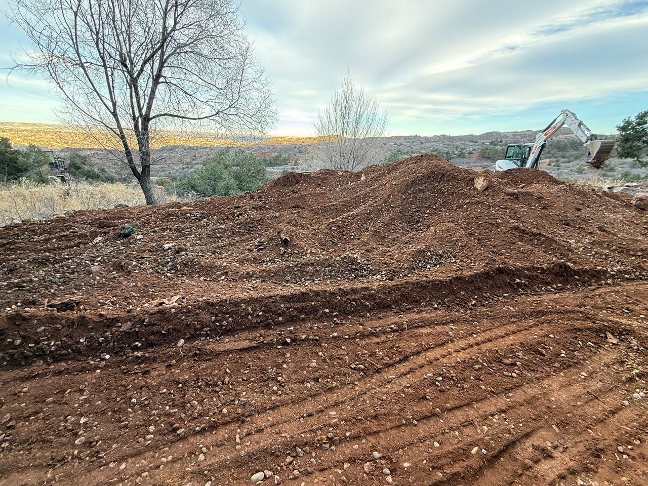 Pile of dirt at a construction site with bare trees and a small house in the background
