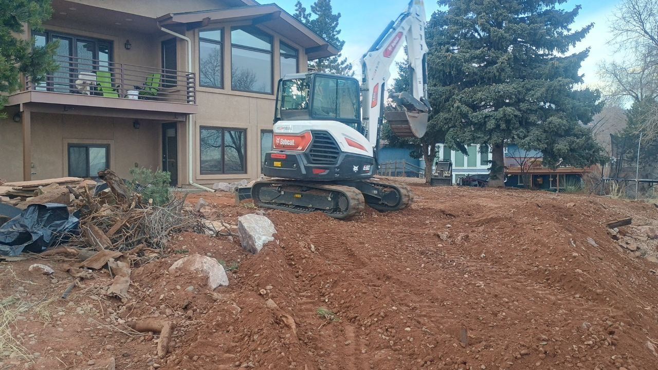 Excavator clearing dirt beside a house under construction, with trees and other homes in the background