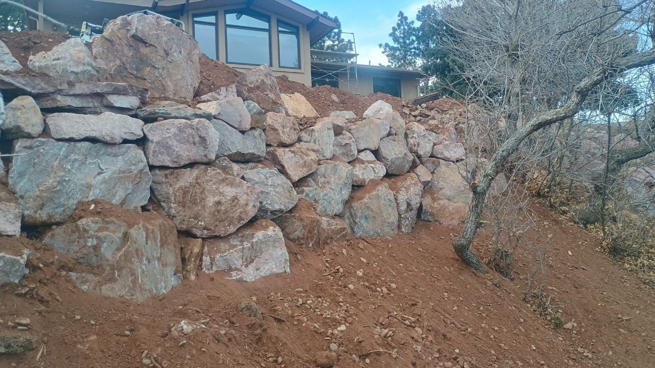 Rock retaining wall on a hillside with a house window and leafless trees above.