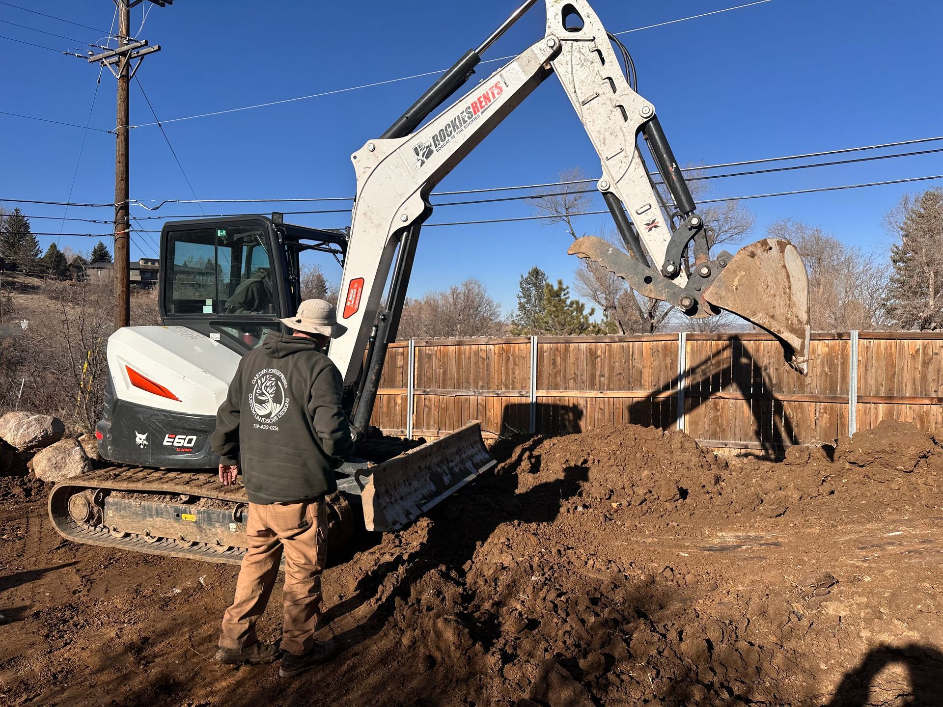 Excavator digging dirt in a backyard, with a worker standing nearby under a clear blue sky.