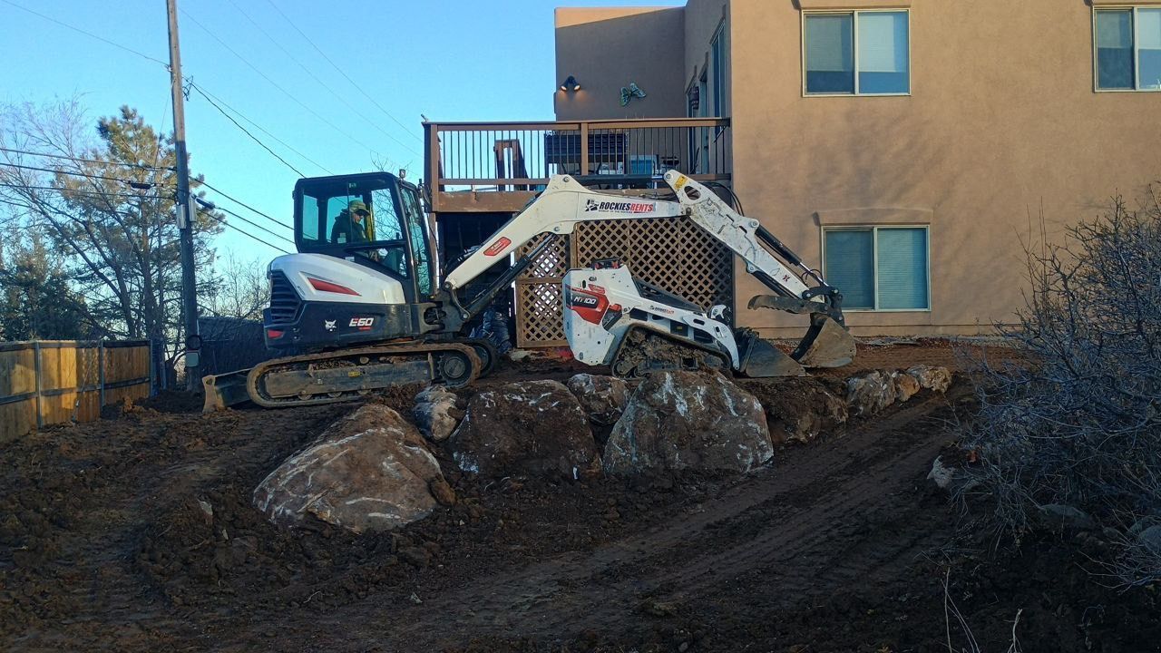 Excavator with a white grapple working on dirt and rocks beside a tan building.
