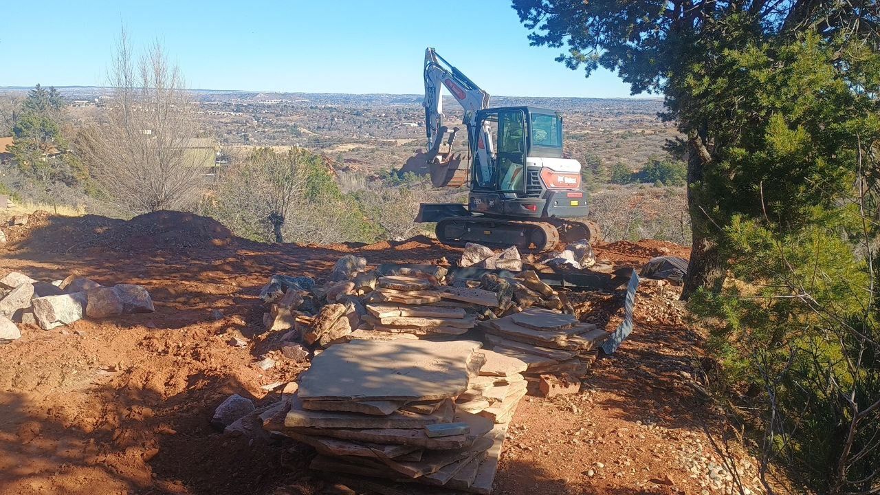 Excavator working on a rocky dirt hillside with trees and a distant landscape in the background
