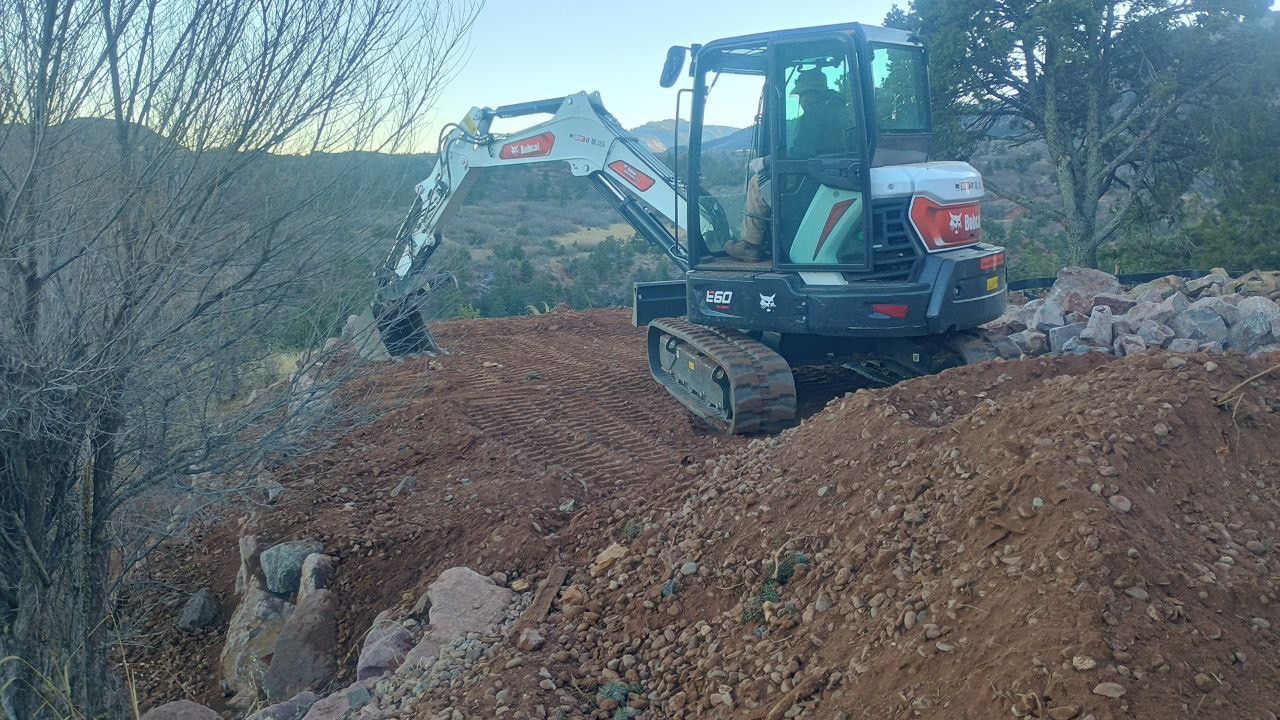 Mini excavator digging on a muddy hillside near leafless trees at dusk