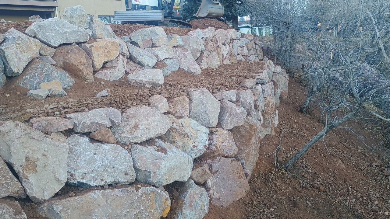 Stone retaining wall beside a dirt path, with bushes and a building in the background.
