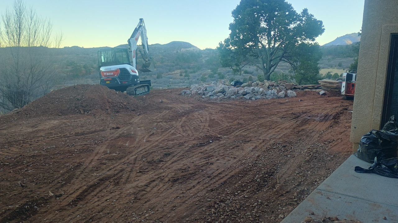 Excavator grading a dirt lot beside a house at dusk, with trees and a hillside in the background