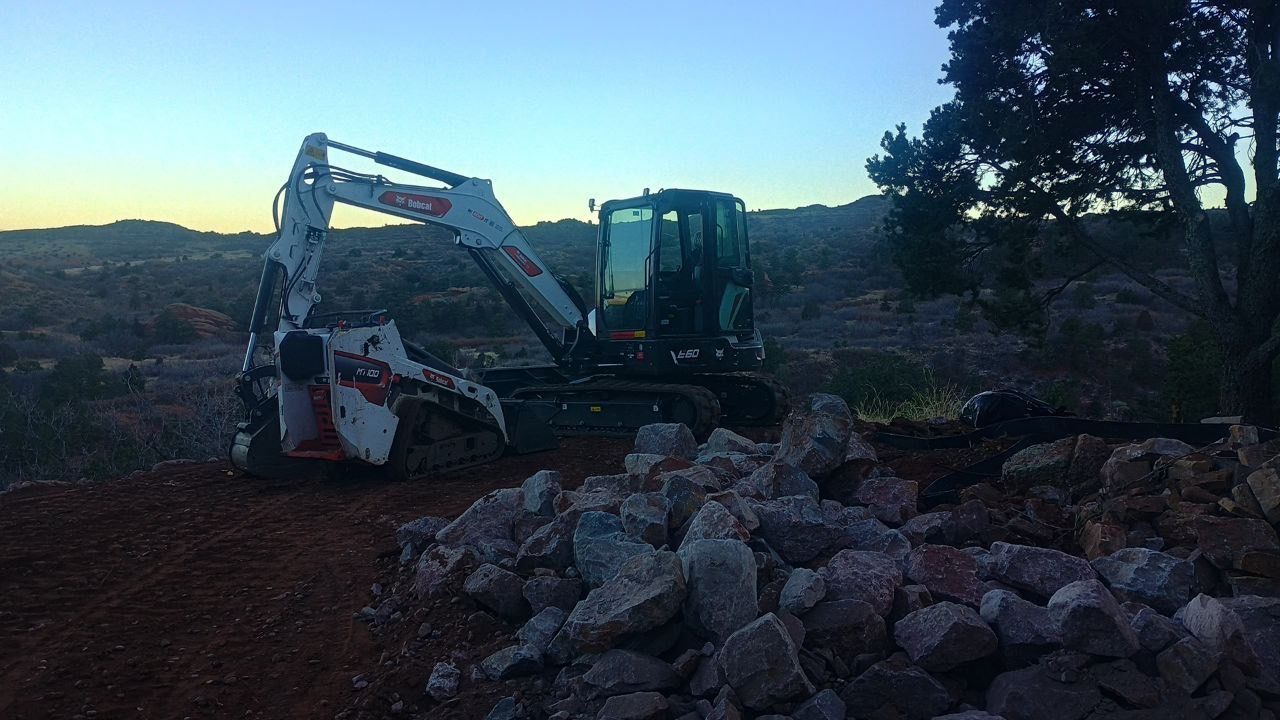 Excavator clearing large rocks at dusk on a dirt construction site, with a blue sky and trees in the background
