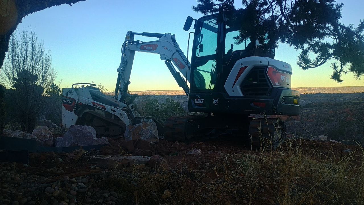 Excavator loading rocks in a field at dusk, silhouetted against a fading blue sky.
