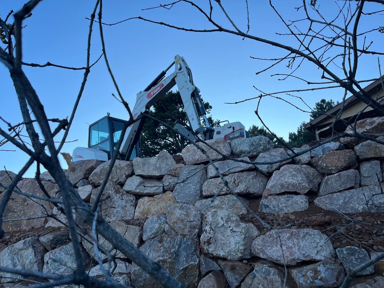 Damaged excavator behind a stacked stone wall with bare tree branches in the foreground under a blue sky