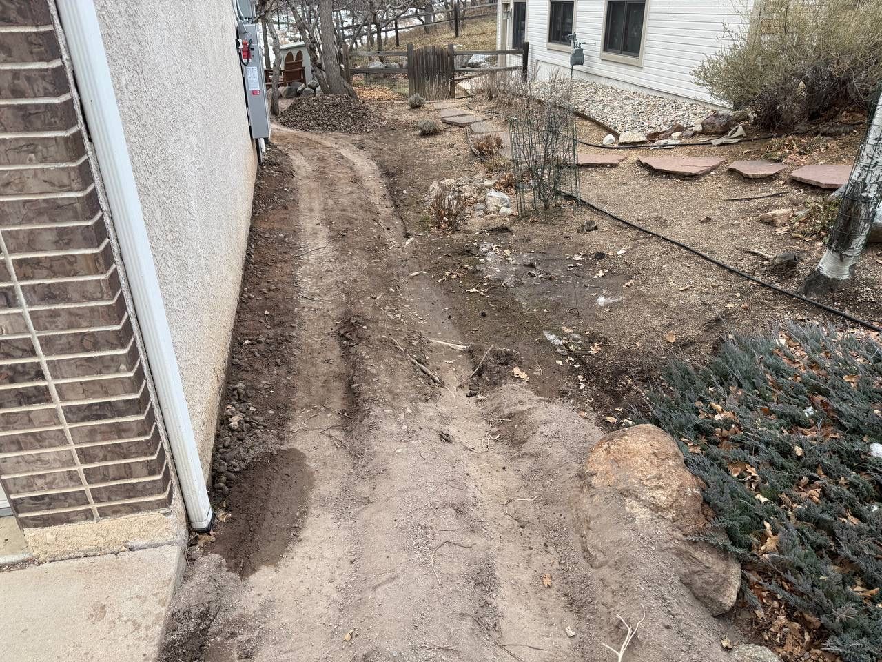 Narrow muddy dirt path beside a house, with wet ground and scattered debris in a backyard.