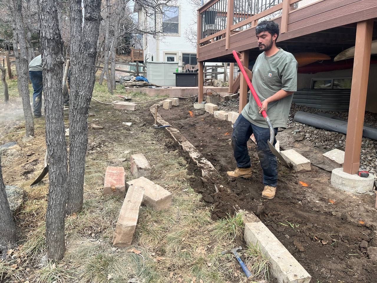 Man in a gray shirt using a red shovel to dig a garden bed beside a wooden deck and trees.