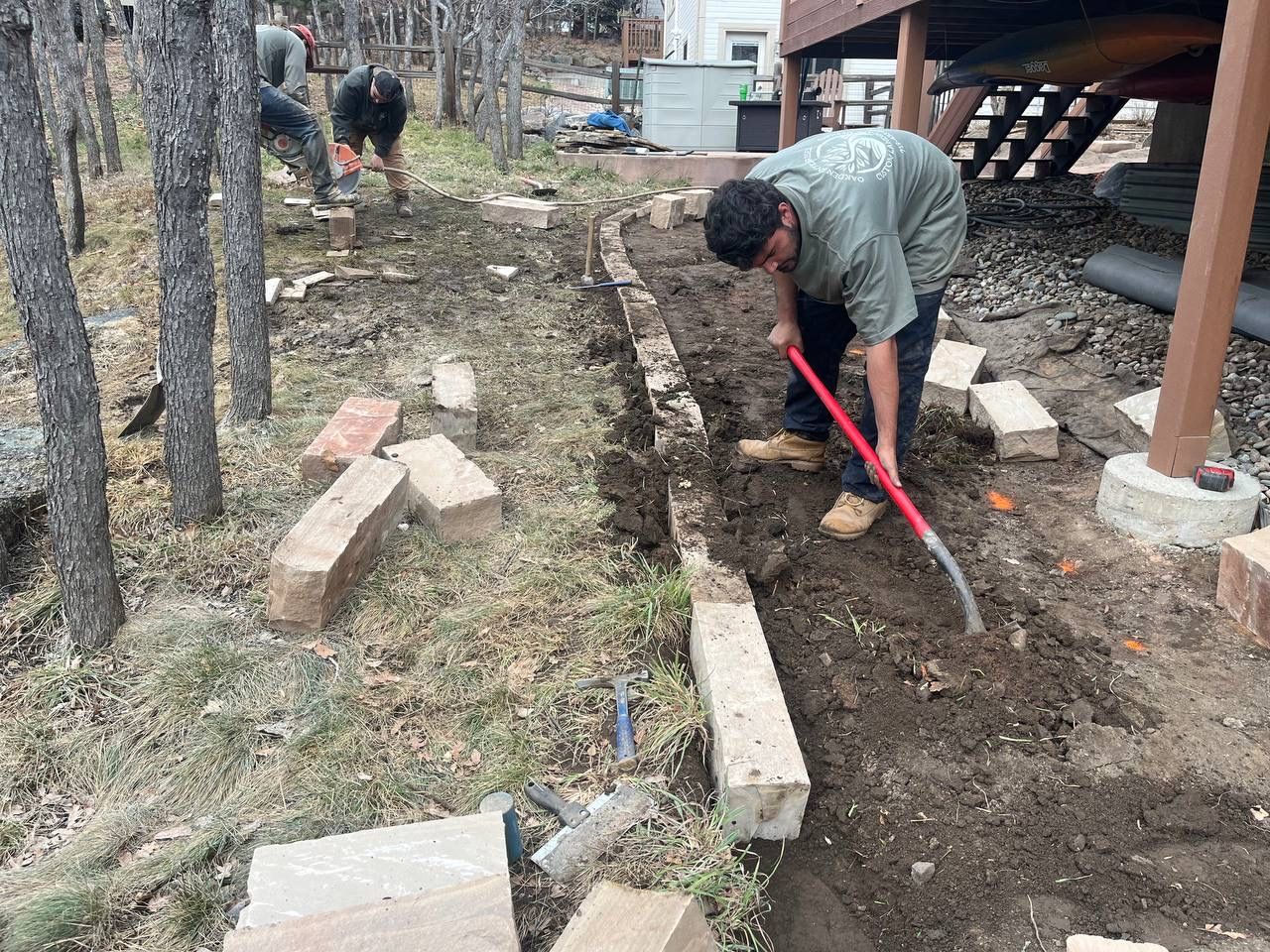 Person digging a trench beside a cabin in a wooded yard with a red shovel