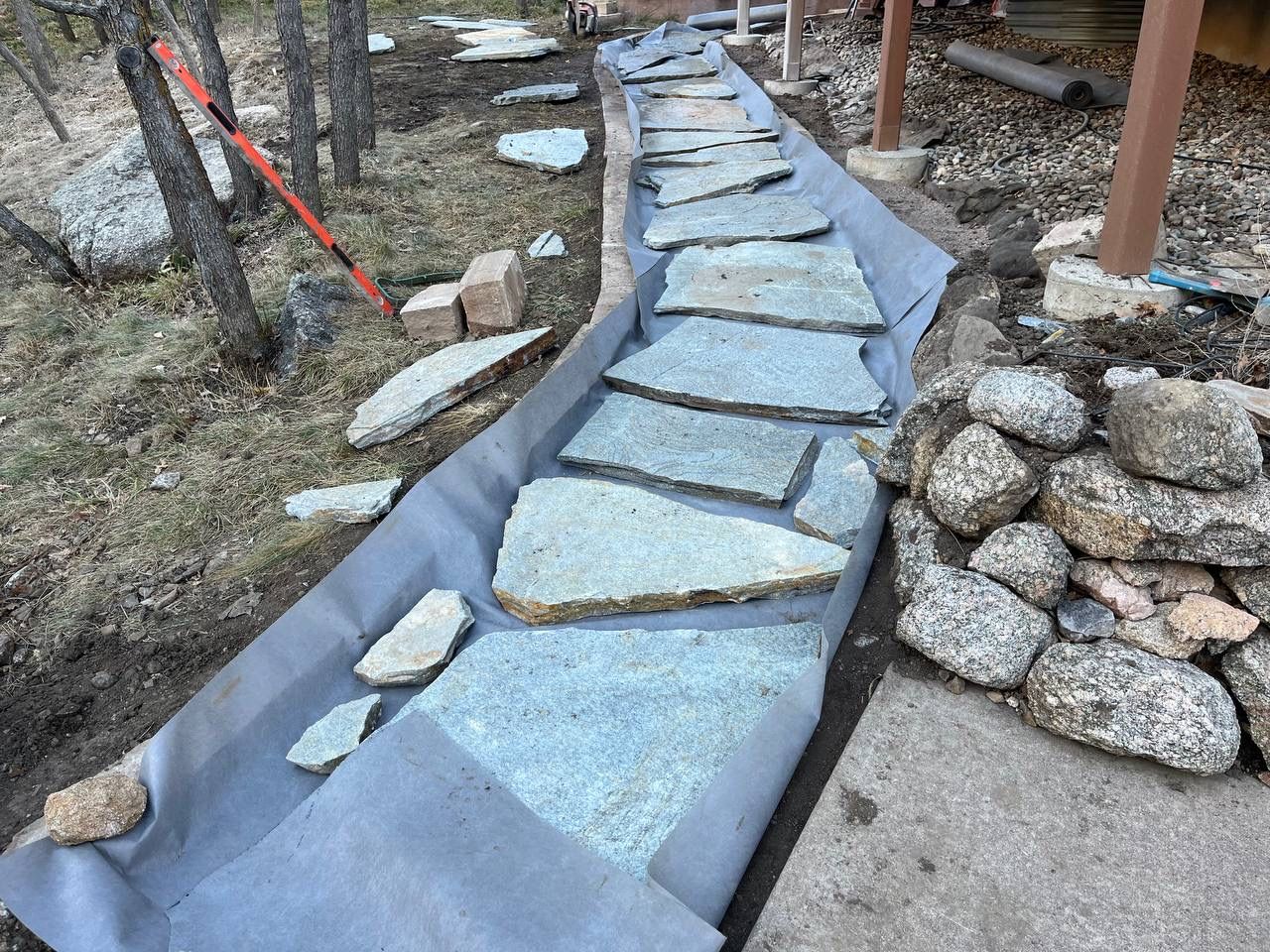 Stone walkway under construction along a rocky slope, with blue underlayment and scattered pavers.